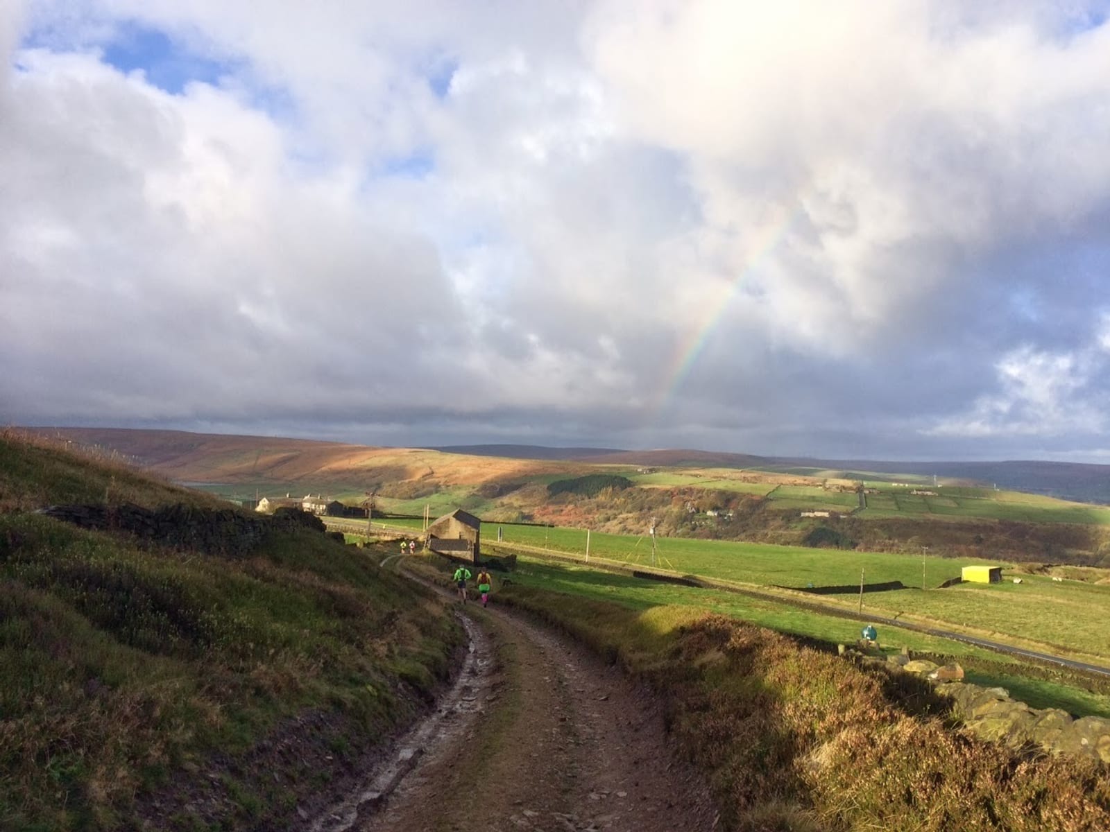 A rural dirt path winds through a green landscape with rolling hills and sparse buildings under a partly cloudy sky. A faint rainbow appears, arcing over the scene, adding a sense of tranquility and beauty to the picturesque countryside.