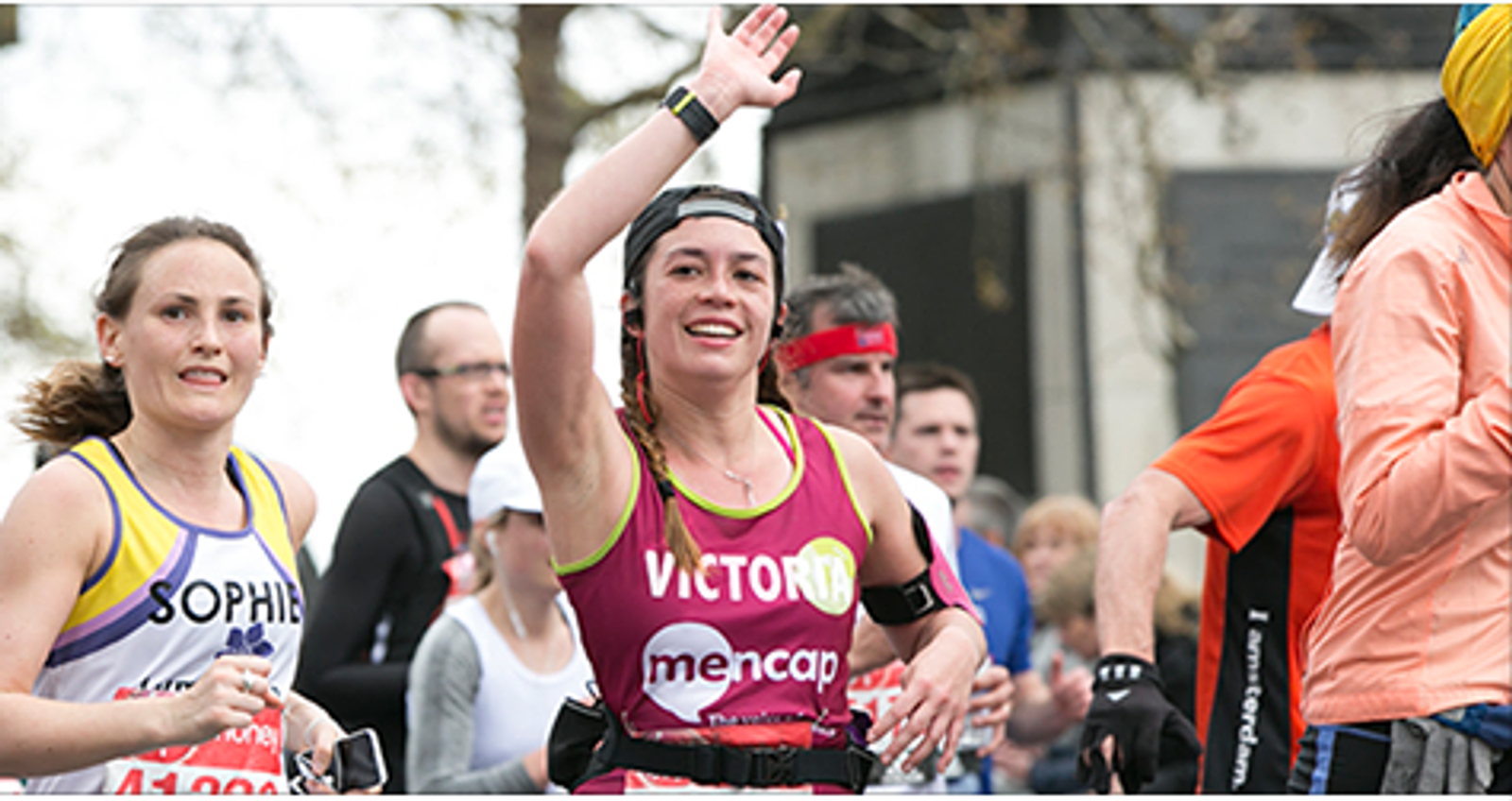Two female runners participate in a race, both wearing athletic gear. The runner in the foreground is smiling, waving, and wearing a cap and a bib reading "VICTOR" and "mencap". The other runner's bib reads "SOPHIE". Several other runners can be seen in the background.