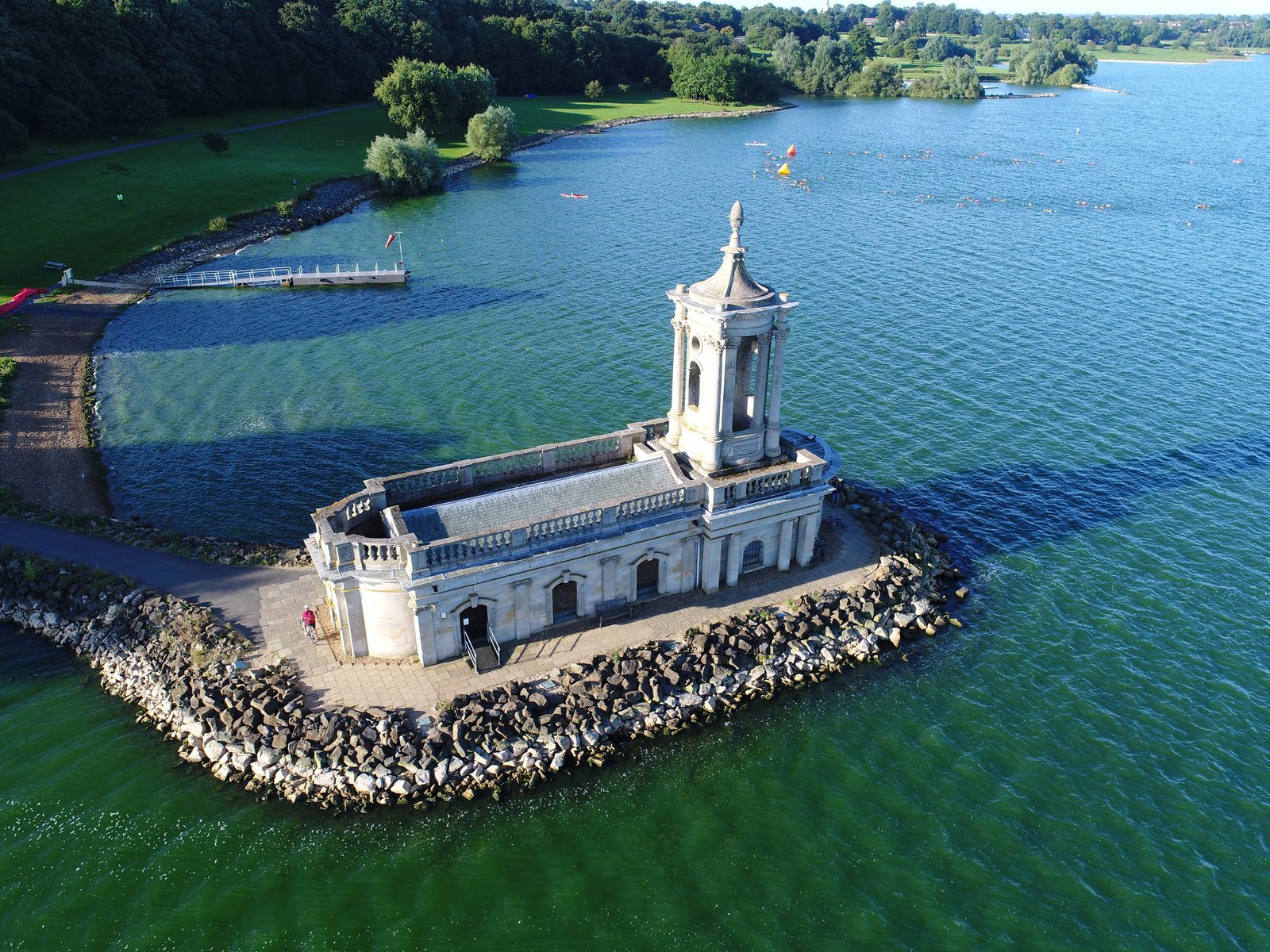 Aerial view of Normanton Church partially surrounded by water, located on the edge of Rutland Water Reservoir. The historic building is bordered by rocks and a path leading to the grassy landscape and treeline in the background on a sunny day.