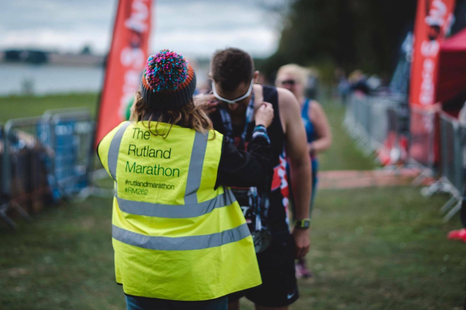 A race volunteer in a bright yellow vest with "The Rutland Marathon" on the back places a medal around the neck of a runner who just finished the marathon. The scene is outdoors with blurred spectators and banners in the background.