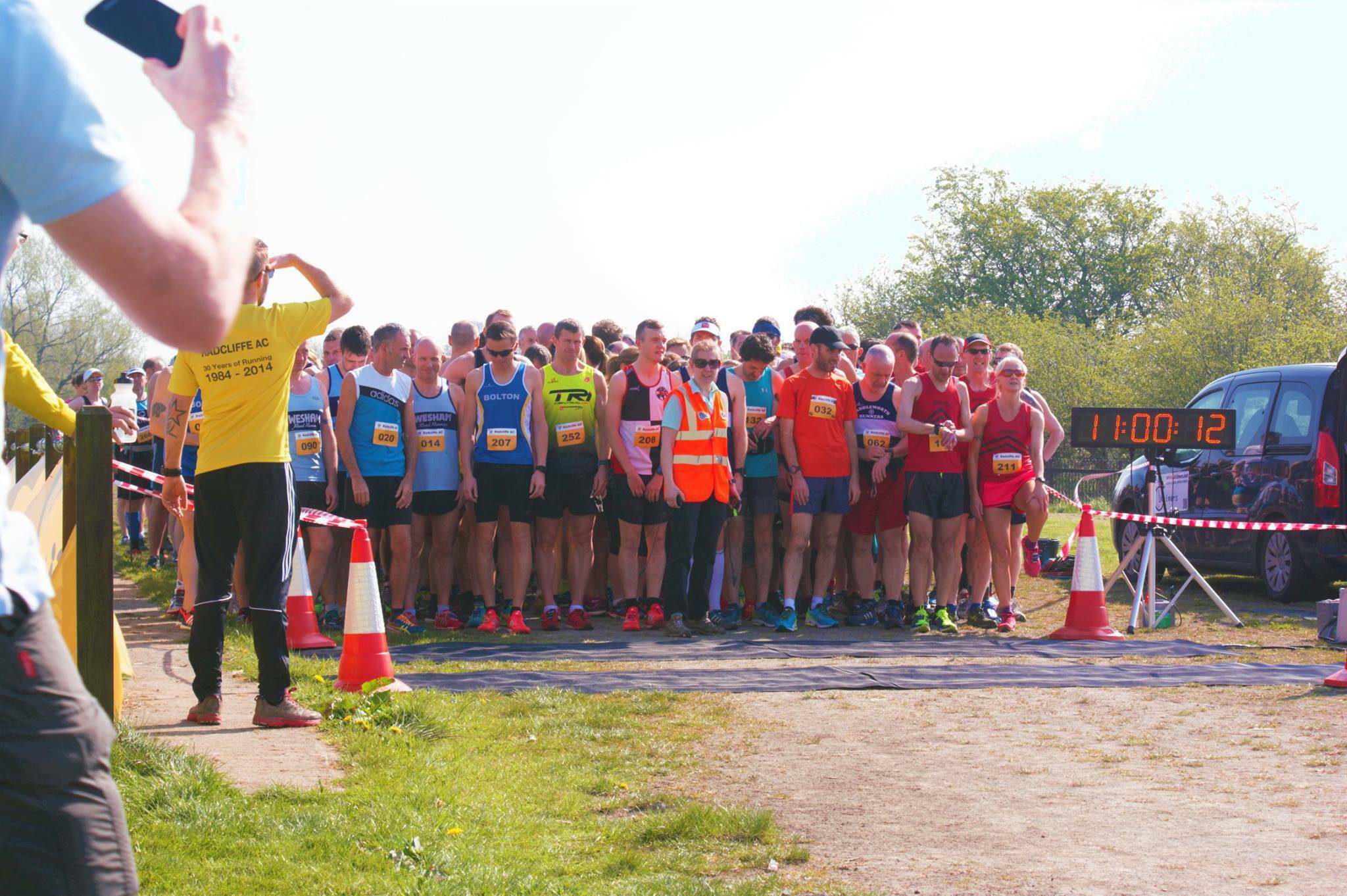 A group of runners is gathered at the starting line of a race on a sunny day. A person in a yellow shirt stands in front, holding a flag. Time clock displays "11:00:12" on the right. Orange cones and tape mark the course boundaries. Trees and grassy area are visible in the background.