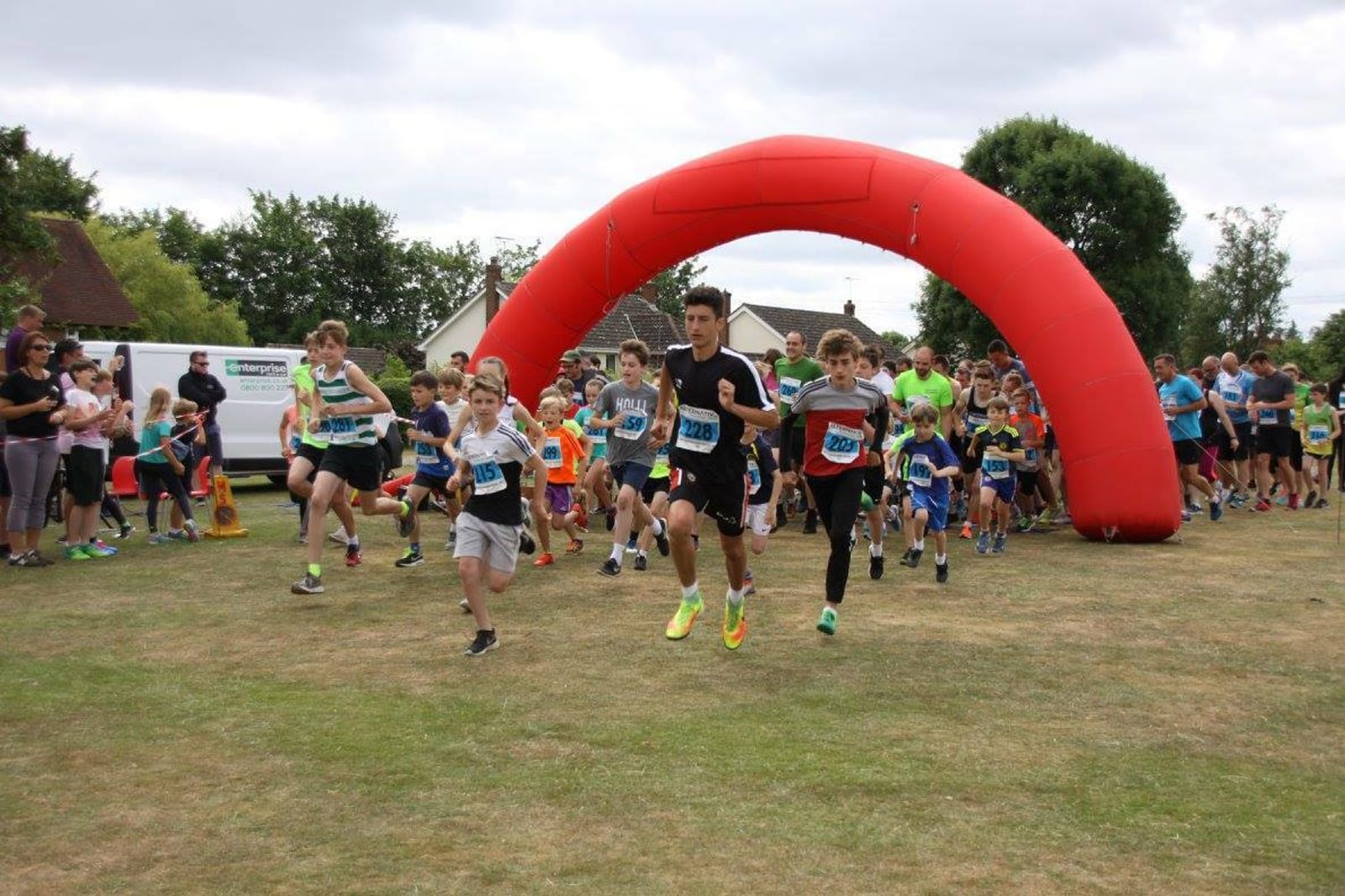 A group of runners of various ages and genders start a race, passing under a large red arch on a grassy field. Spectators and other participants stand on the sidelines, cheering them on. It’s a sunny day with partly cloudy skies.