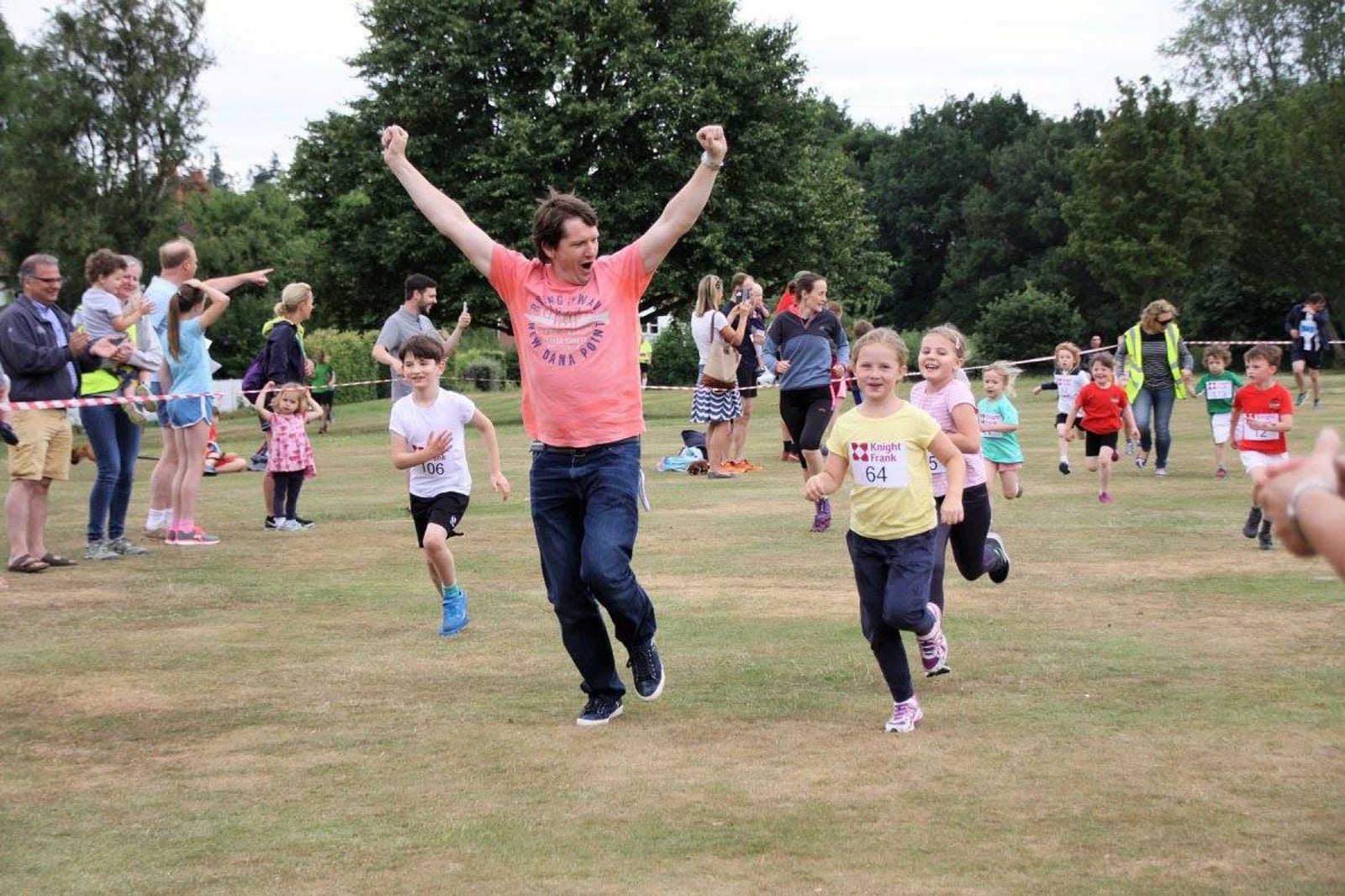 A man with raised arms is running alongside children participating in a race. Several adults and children are watching and cheering from the sidelines. The children are wearing numbered bibs, and the scene takes place on a grassy field with trees in the background.