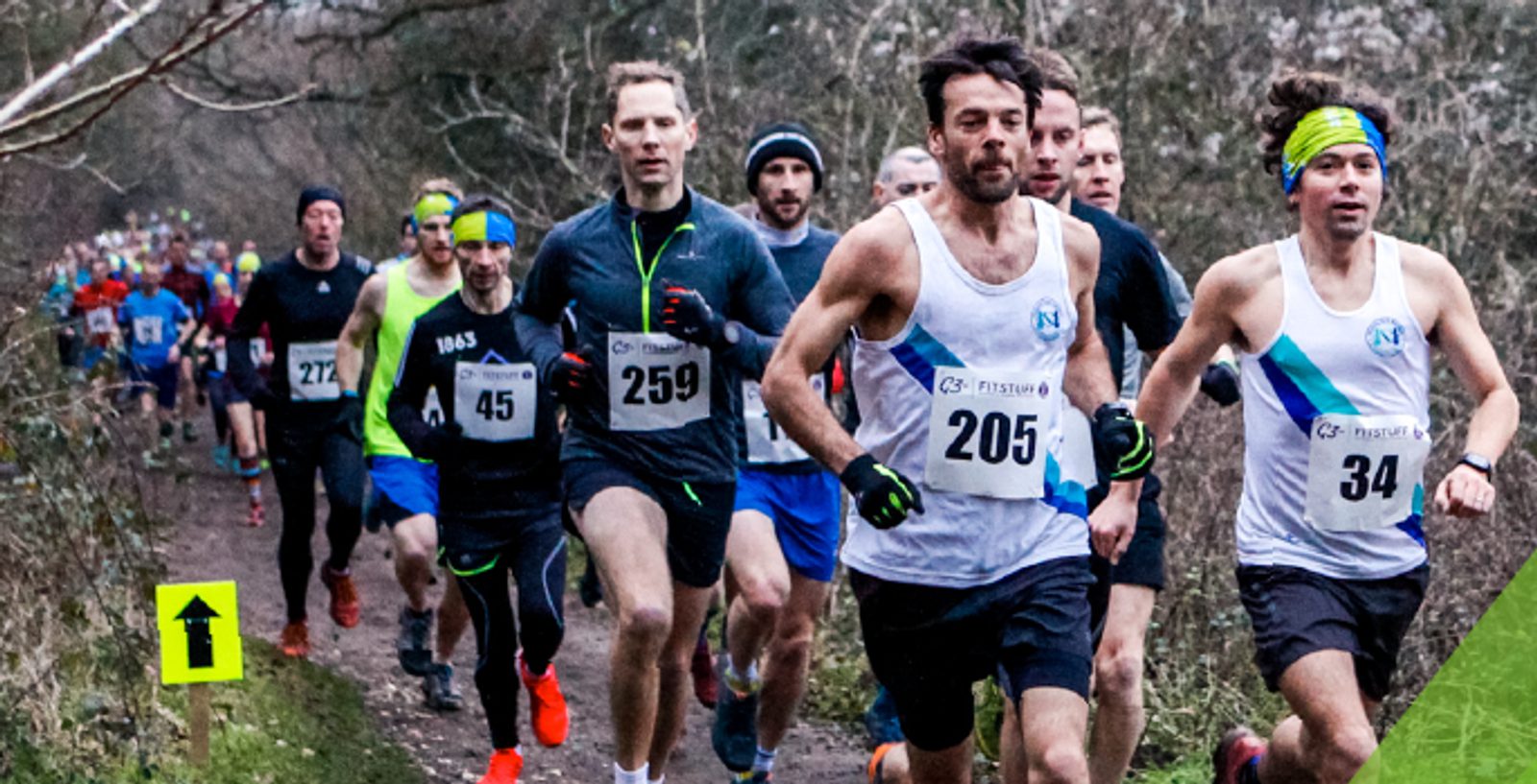 A group of runners on a forest trail, participating in a race. They wear numbered bibs and various athletic outfits, including tank tops, long-sleeve shirts, and headbands. A green arrow sign points them in the correct direction. Trees and branches line the path.