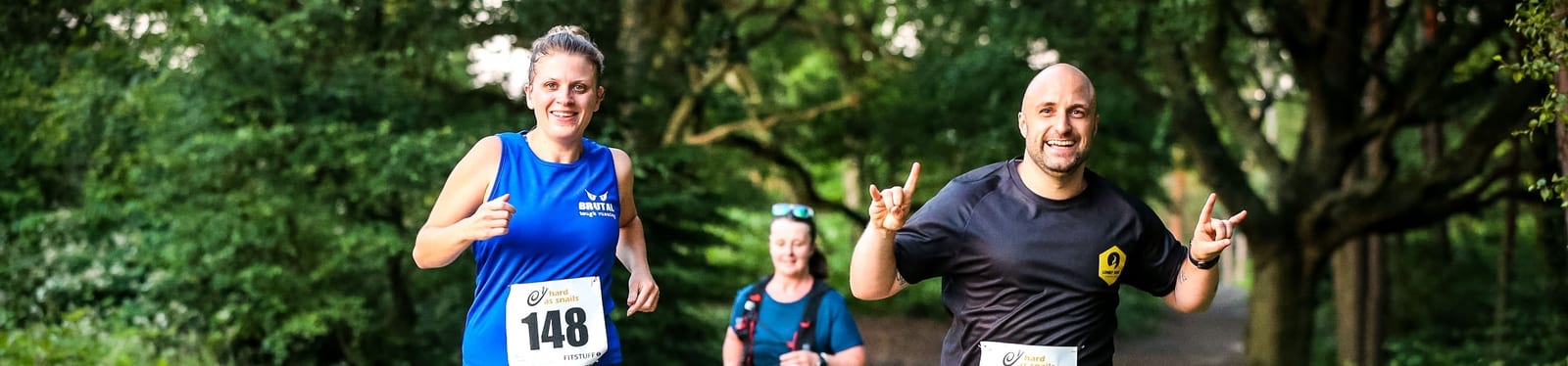 Three people running in a park, smiling and wearing numbered bibs. The woman on the left is in a blue tank top, the man on the right is in a black t-shirt, and the other runner behind them is in a blue shirt. Tall trees and greenery fill the background.