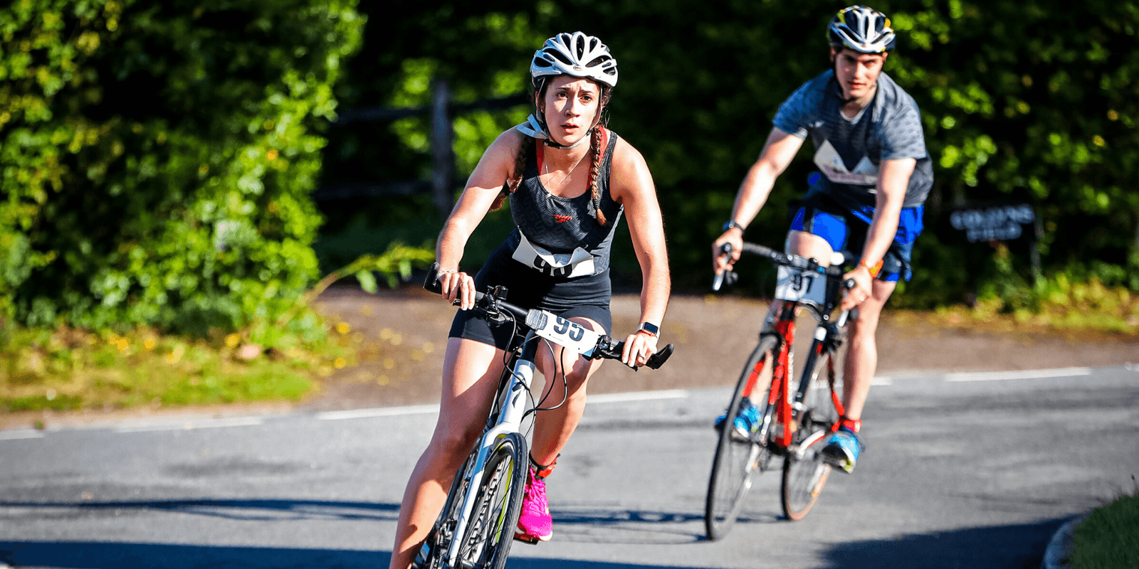 Two cyclists participate in a race, riding road bikes around a bend. The foreground cyclist, wearing a white helmet, black outfit, and pink shoes, appears focused. The background cyclist, dressed in a gray shirt and blue shorts, follows closely behind. Greenery surrounds the road.