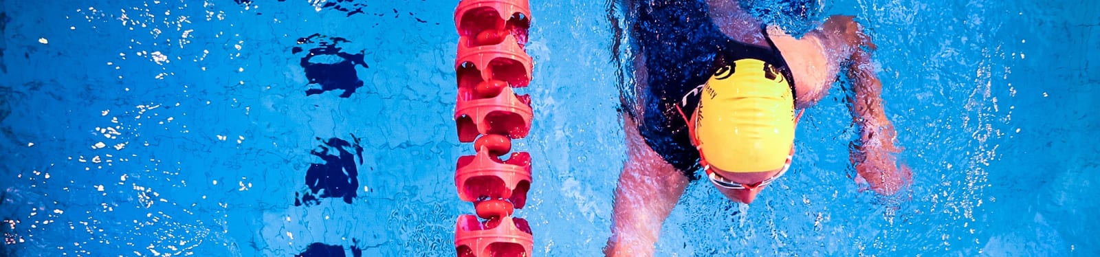 A swimmer in a yellow cap and dark swimsuit competes in a swimming pool, near a lane divider with red buoys. The swimmer is captured mid-stroke as they move through the blue water, creating splashes.