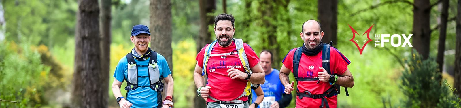 Three men are running through a forested trail, visibly enjoying the run. One man wears a blue shirt and two others wear red shirts, with all carrying hydration packs. The logo and text "The Fox" appears on the right side of the image. Trees and greenery form the backdrop.