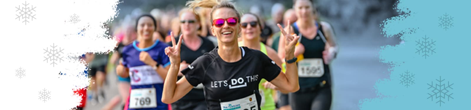 A group of enthusiastic runners participating in a race. The central figure, a woman in a black T-shirt that says "LET'S DO THIS," is smiling and making a hand gesture with both hands. She is wearing pink sunglasses, and other runners are visible in the background.