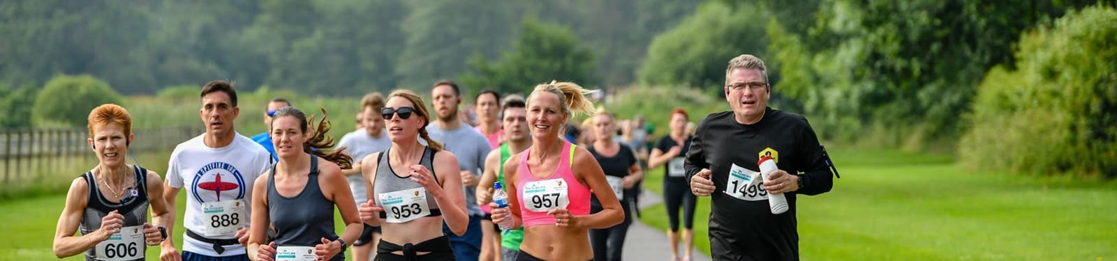 A group of runners participating in an outdoor race. They are running along a paved path surrounded by lush green scenery. The participants wear race bibs with numbers and appear focused on the race. The background features trees and grass.