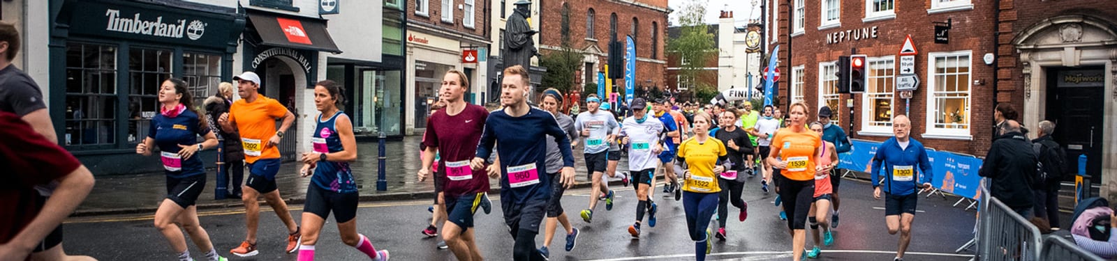 A diverse group of runners wearing brightly colored athletic gear participate in a road race through a town with historic buildings and shopfronts, including a Timberland store. Spectators and organizers are seen along the sides, with barriers set up on the wet street.