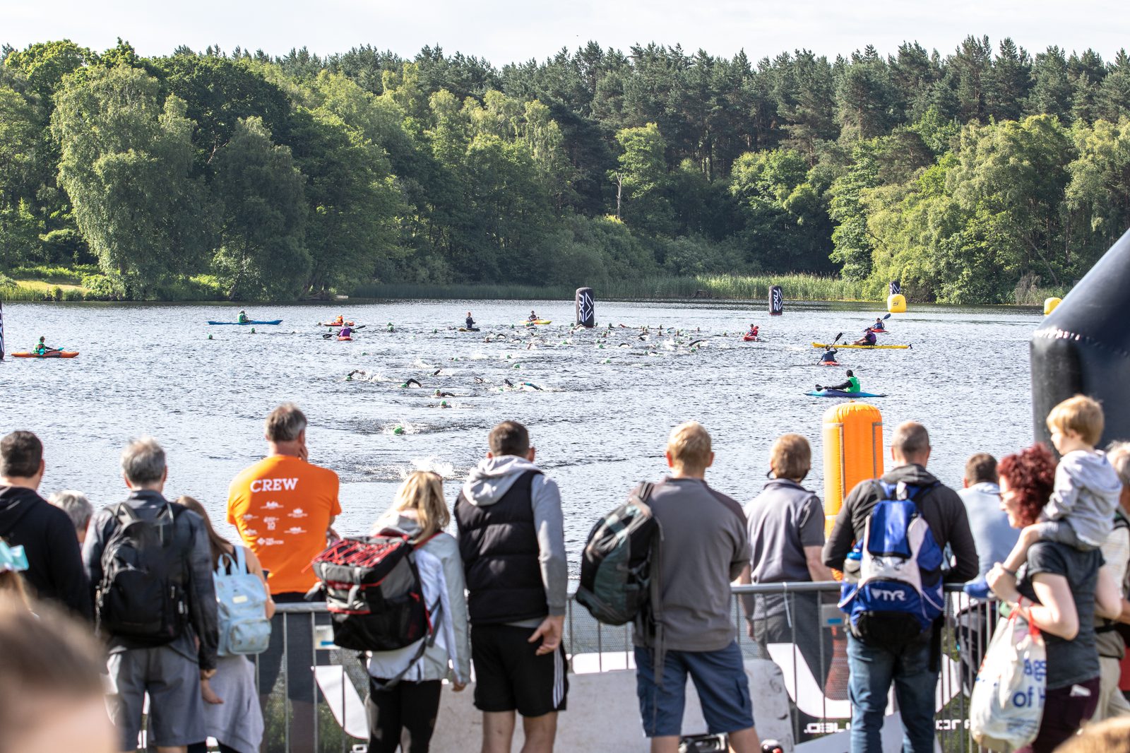 A crowd watches swimmers participating in an open water race in a lake surrounded by trees. Kayaks and buoys are visible in the water, guiding the swimmers. Some spectators wear backpacks and jackets, while others have strollers and children.