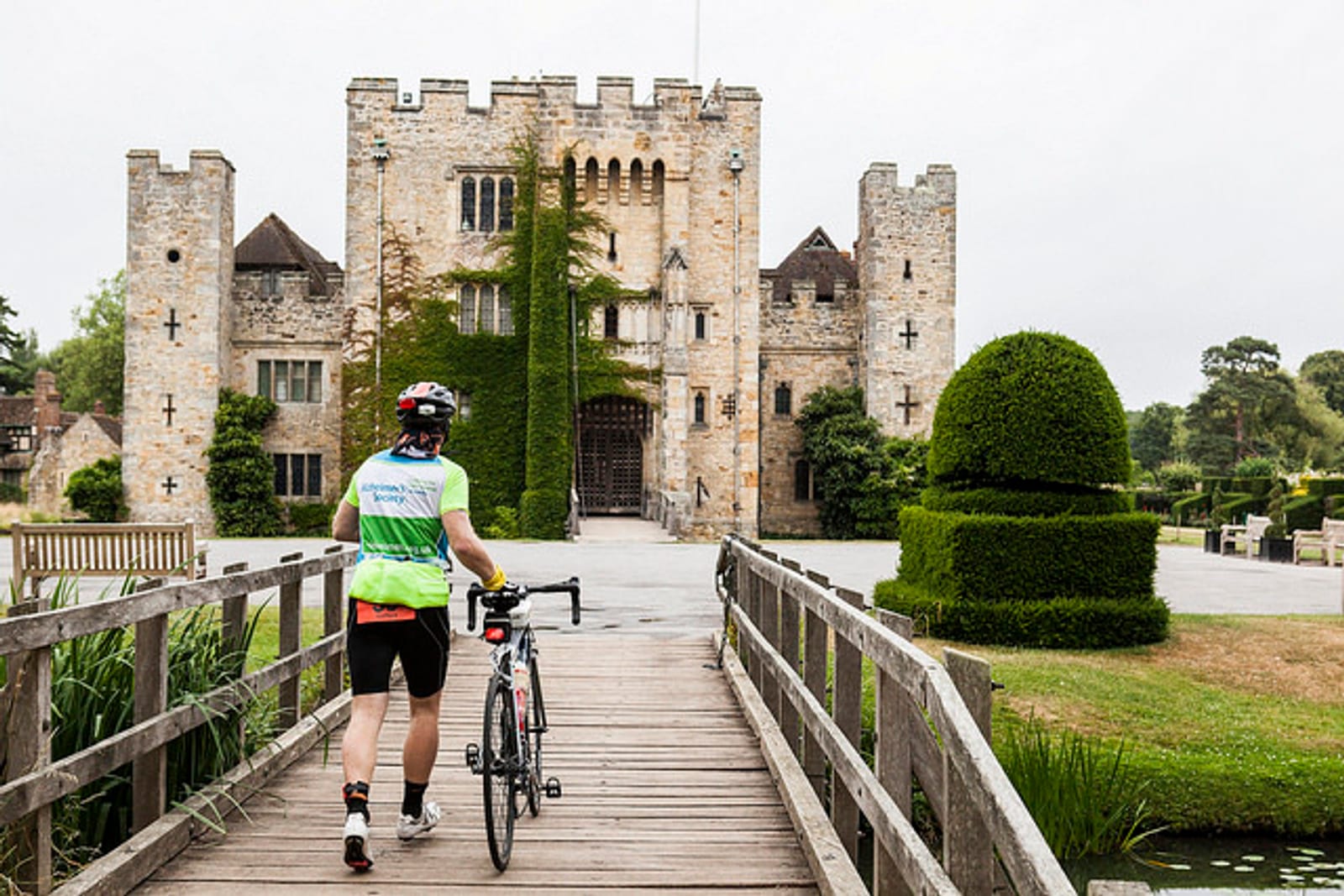 A cyclist wearing a helmet, cycling clothing, and holding a bicycle walks across a wooden bridge toward a large, historic stone castle with a manicured garden. The sky is overcast, and the setting exudes a tranquil, historic atmosphere.
