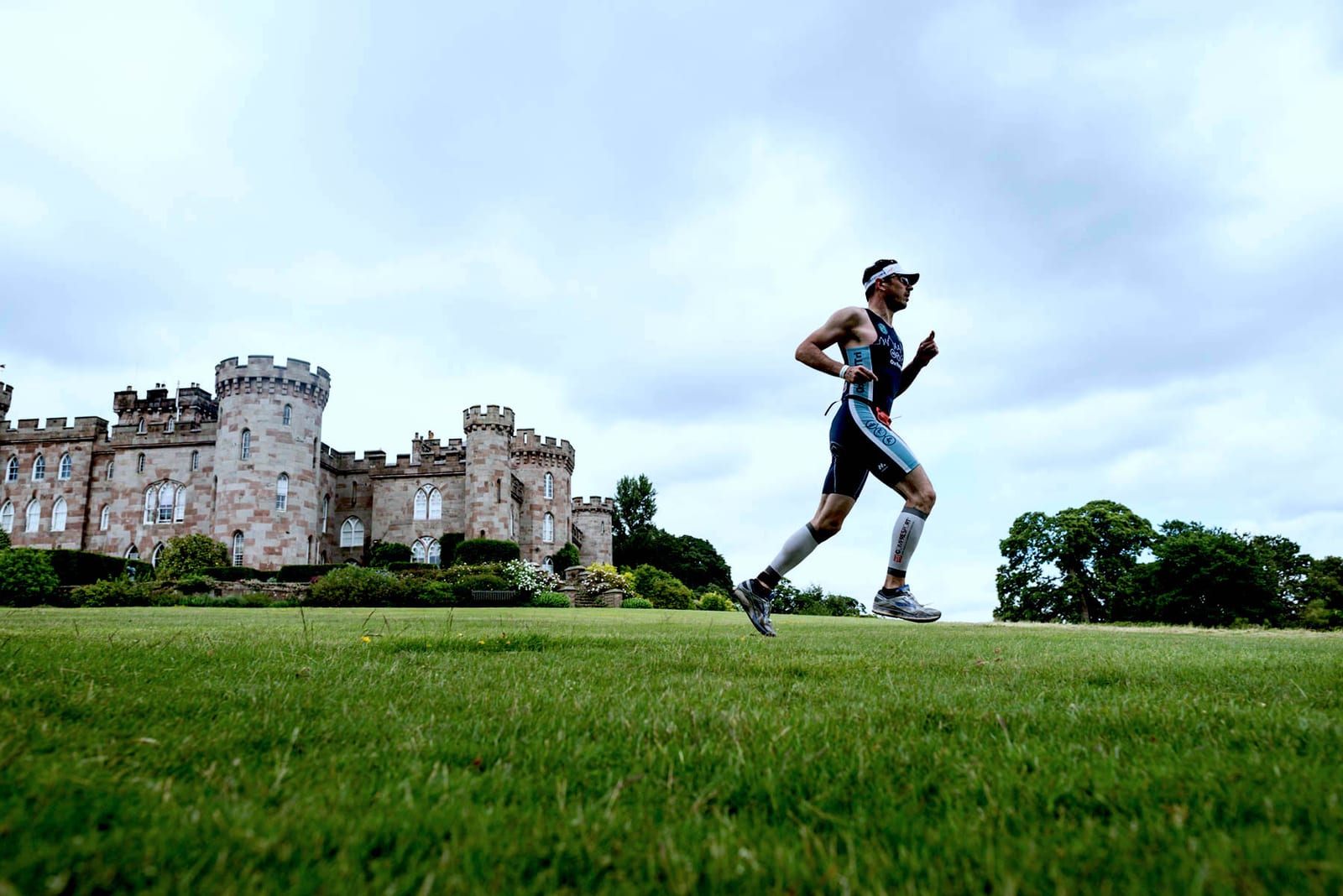 A person in athletic gear and sunglasses runs on a grassy field in front of a historic stone castle with turrets and battlements, under a cloudy sky. Trees and shrubs are visible in the background.