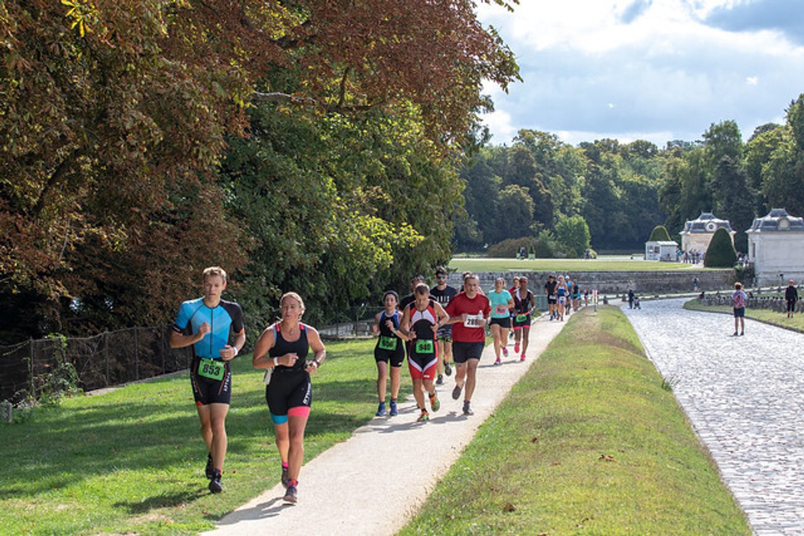 A group of people jogging along a riverside path lined with trees and greenery on a sunny day. The runners are wearing athletic clothing and numbered bibs, indicating participation in a race or fun run event. The path is adjacent to a cobblestone walkway.