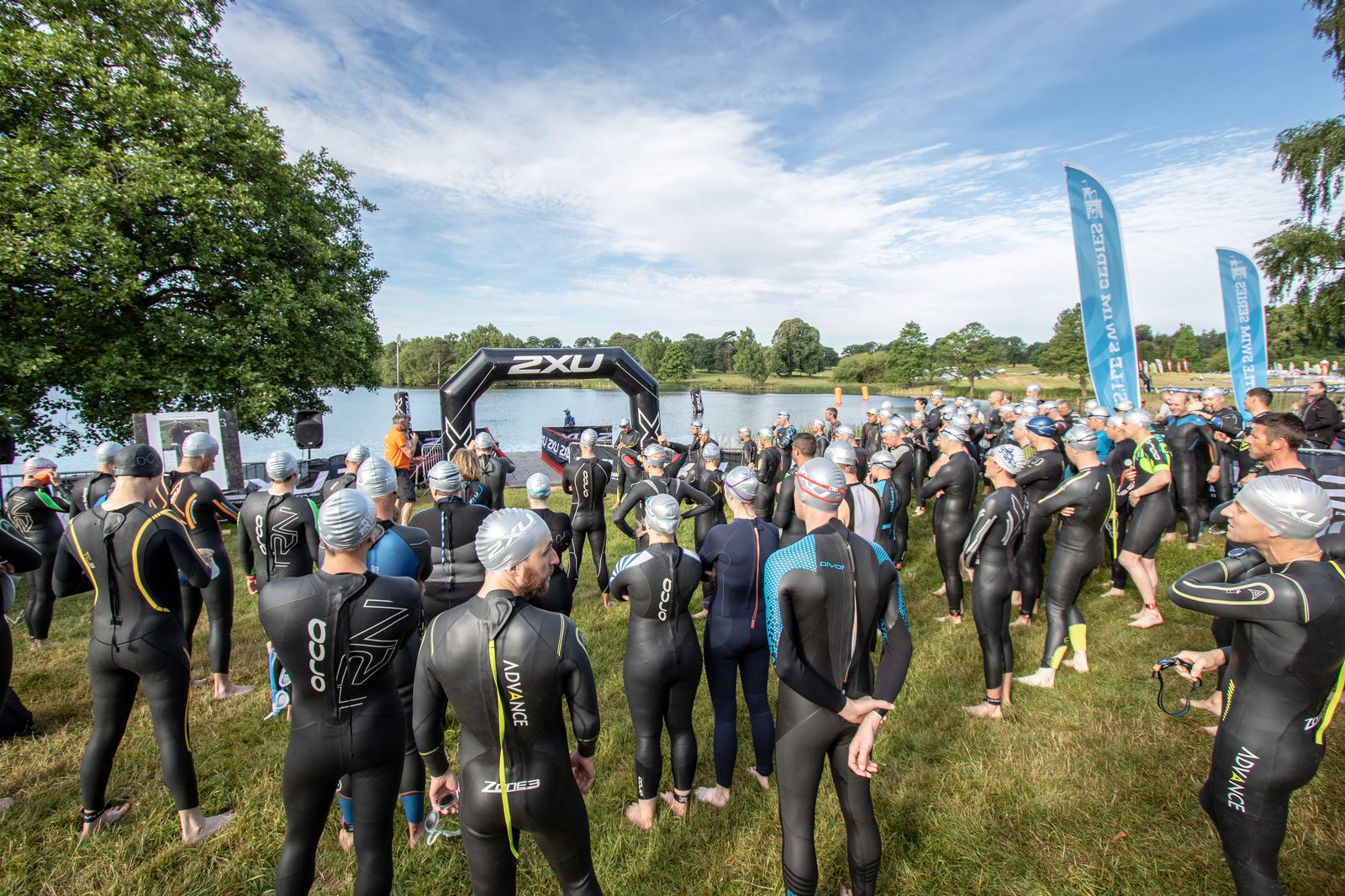 A large group of triathletes in wetsuits and swim caps stand on a grassy area near a lake, listening to instructions before a race. An inflatable archway displaying "2XU" is set up at the water's edge, and trees and a blue sky with clouds are visible in the background.
