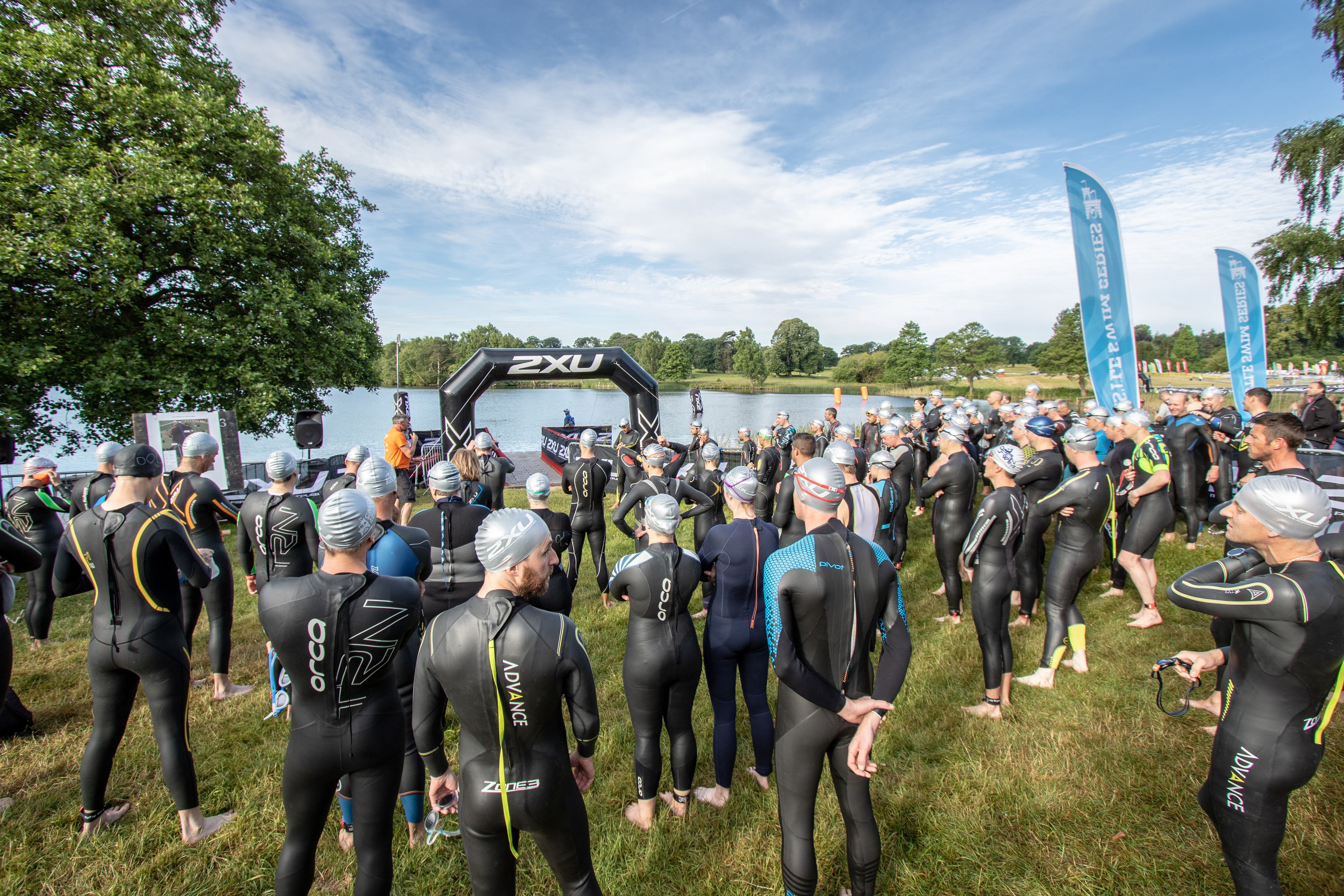 A large group of triathletes, all wearing wetsuits and swim caps, gather near a lake under a clear sky for the start of a race. They stand in front of an inflatable archway and listen to instructions. Trees and greenery are visible in the background.