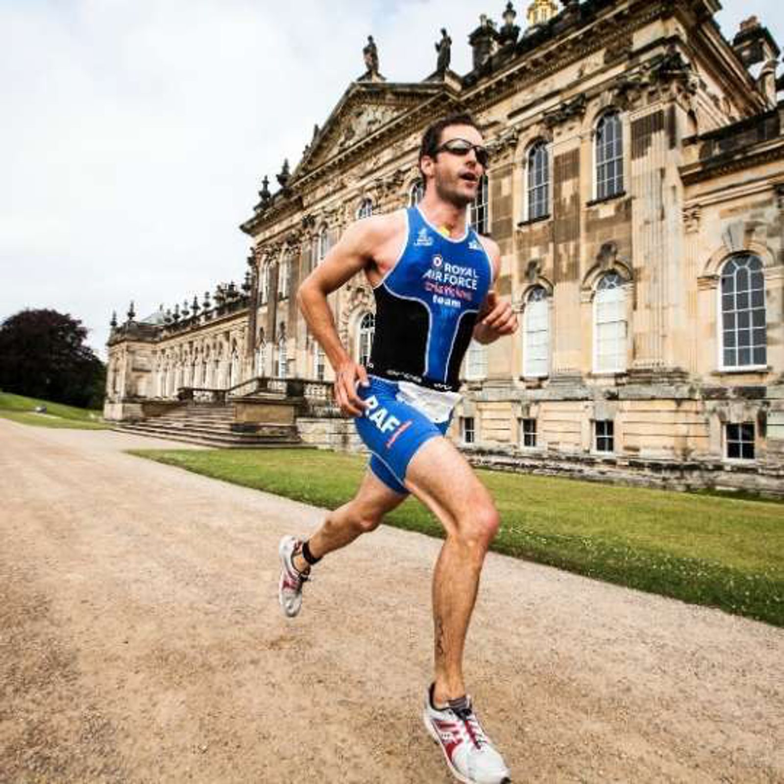 A man in athletic clothing runs on a gravel path past a large historic building, with "Royal Air Force" printed on his outfit. He wears sunglasses and appears focused on his run. The sky is clear, and the scene suggests a race or marathon event.