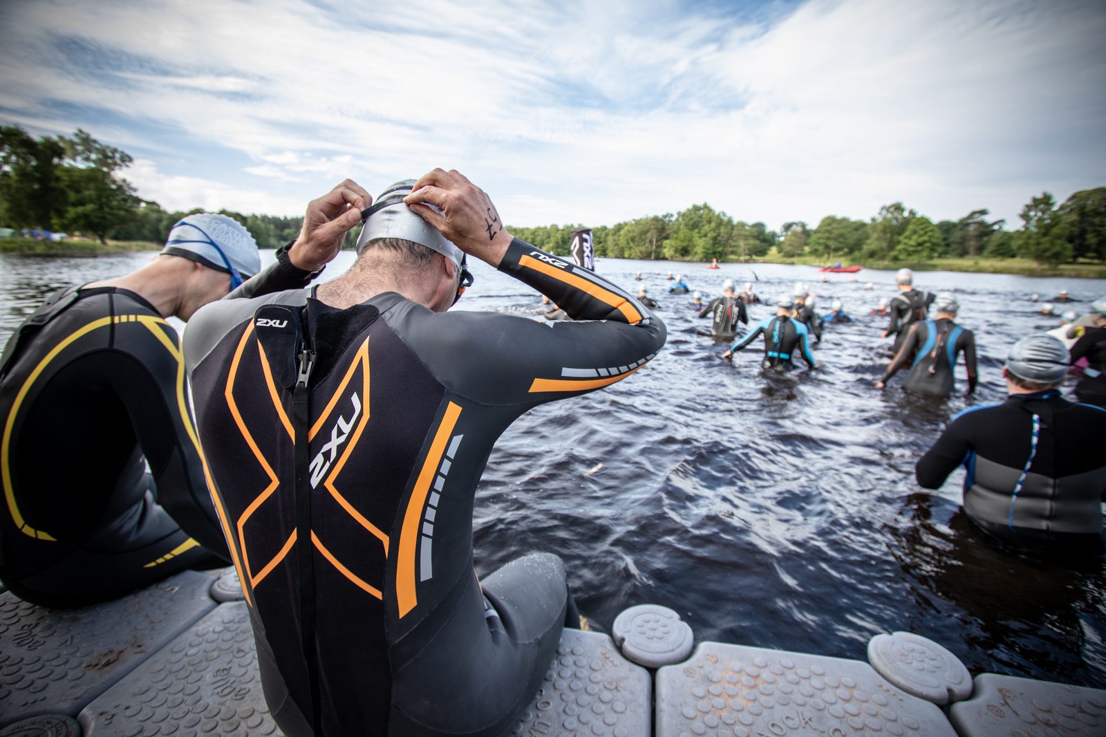 A group of swimmers wearing wetsuits and swim caps prepare for a race in an outdoor body of water. One swimmer sits on a dock adjusting their swim cap, while others stand in the water waiting for the event to start. Trees and a cloudy sky are in the background.