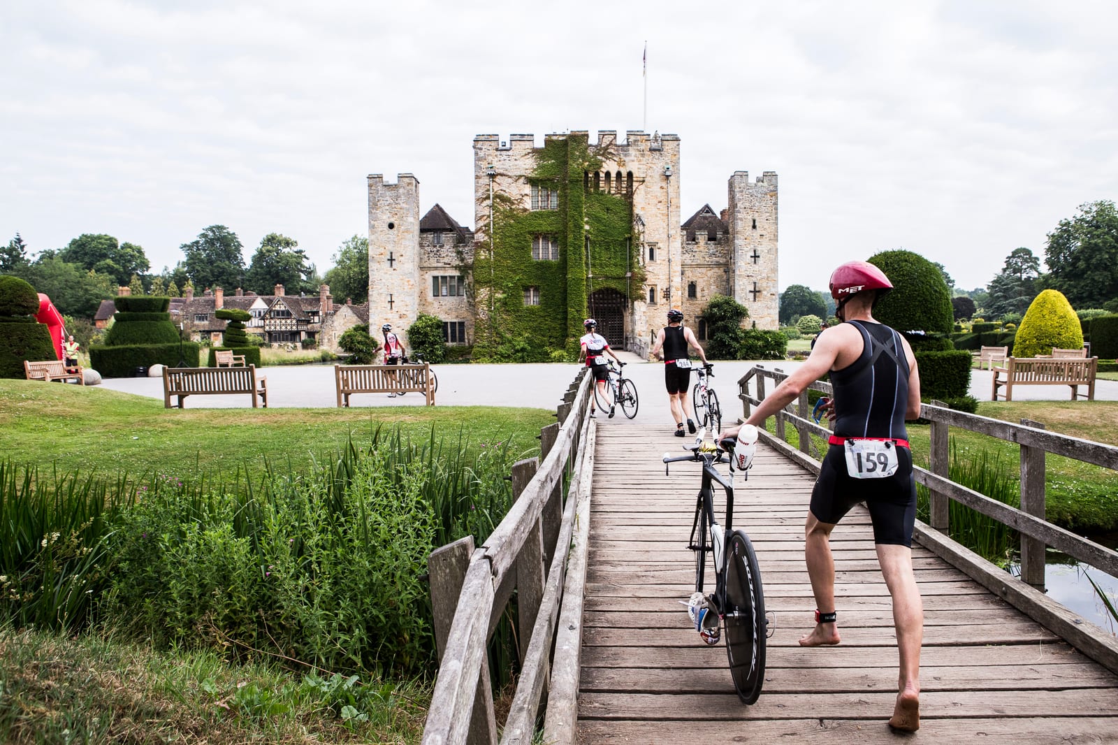 A group of athletes in sports attire cross a wooden bridge with their bicycles towards a castle with a stone facade and turrets, partially covered in ivy. The scene is set against a backdrop of lush green lawns, neatly trimmed hedges, and a cloudy sky.