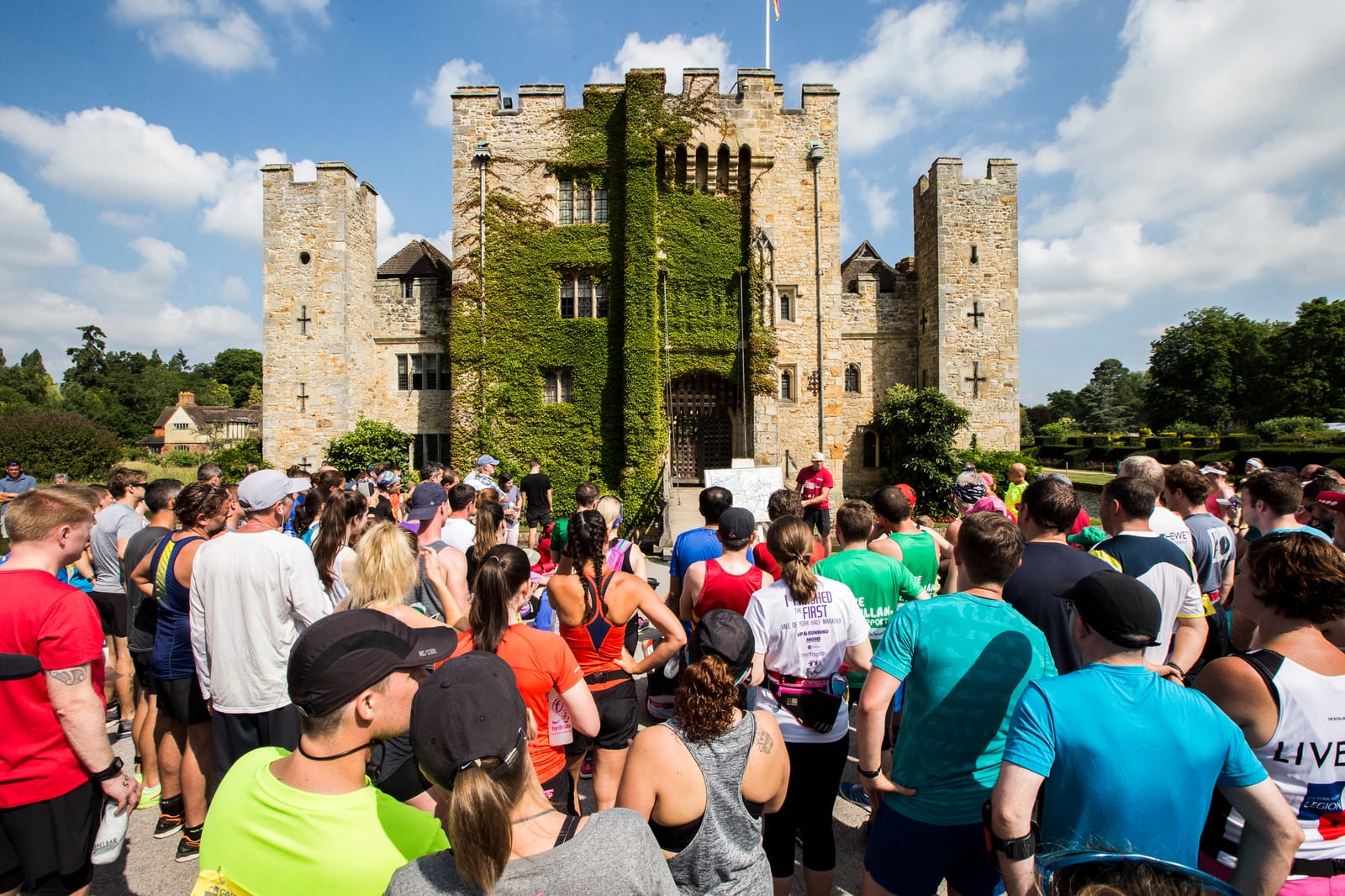 A crowd of people dressed in athletic gear are gathered in front of an ancient stone castle covered with ivy. The sky is partly cloudy, and the mood is lively as the group prepares for an event or run on a sunny day.
