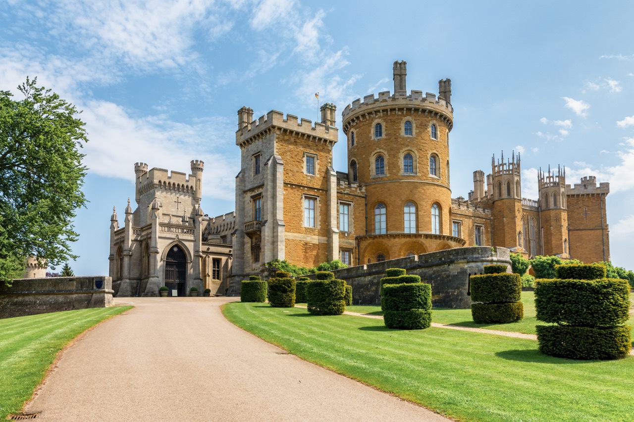 A grand castle with multiple towers and spires stands under a partly cloudy blue sky. The castle is made of stone and features intricate architectural details. A well-maintained garden with trimmed hedges and a winding path leads to the entrance.