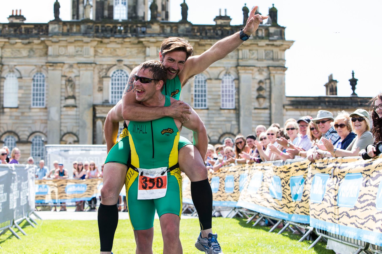 Two male athletes in green and yellow triathlon suits celebrate at a race finish line, with one jumping onto the other's back. They are smiling and cheering, surrounded by applauding onlookers. A historic building is visible in the background under a sunny sky.