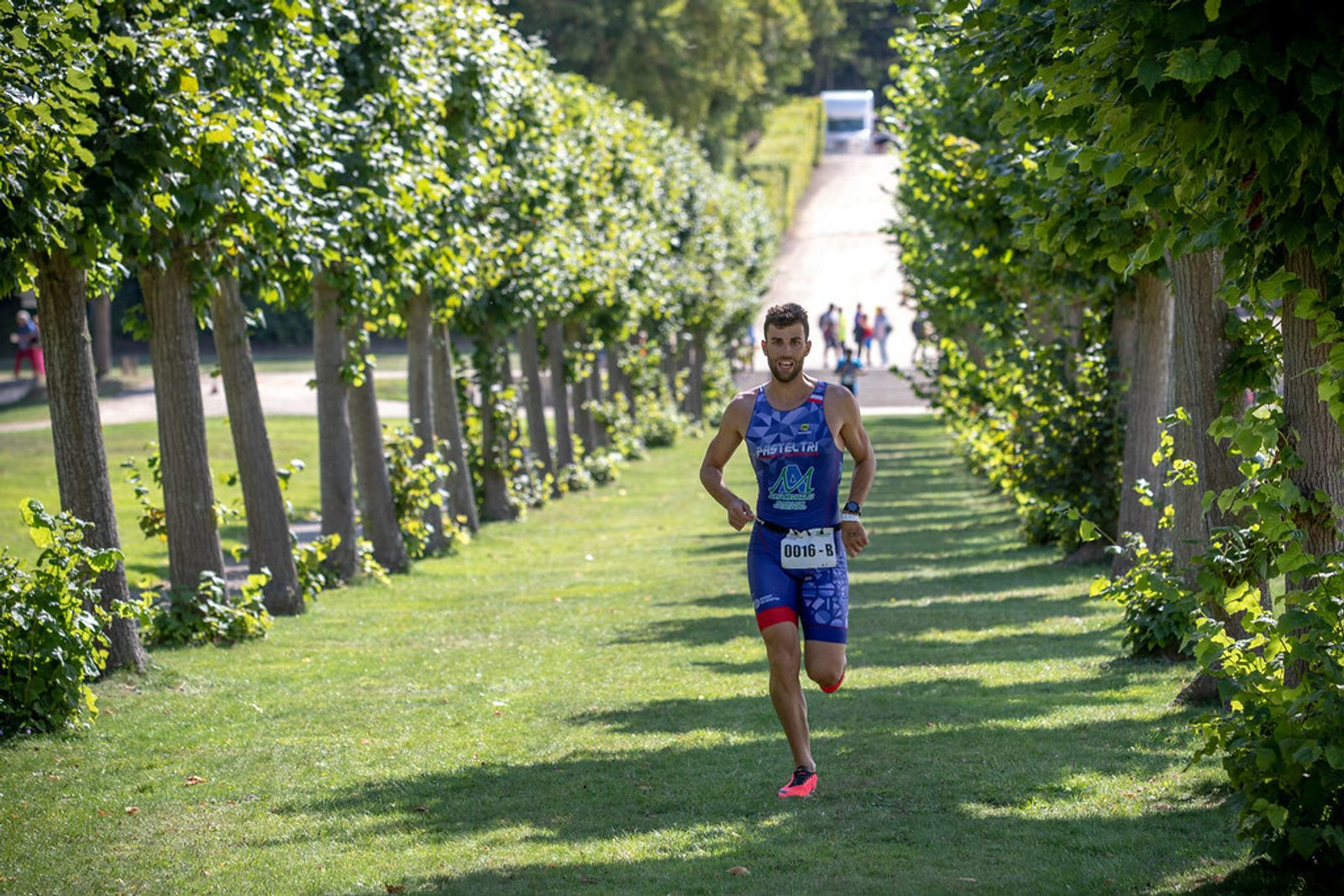 A man in athletic gear runs on a tree-lined path during a race. Sunlight filters through the trees, casting shadows on the grass. Other runners can be seen in the distance. The setting appears to be in a park or garden with lush greenery.