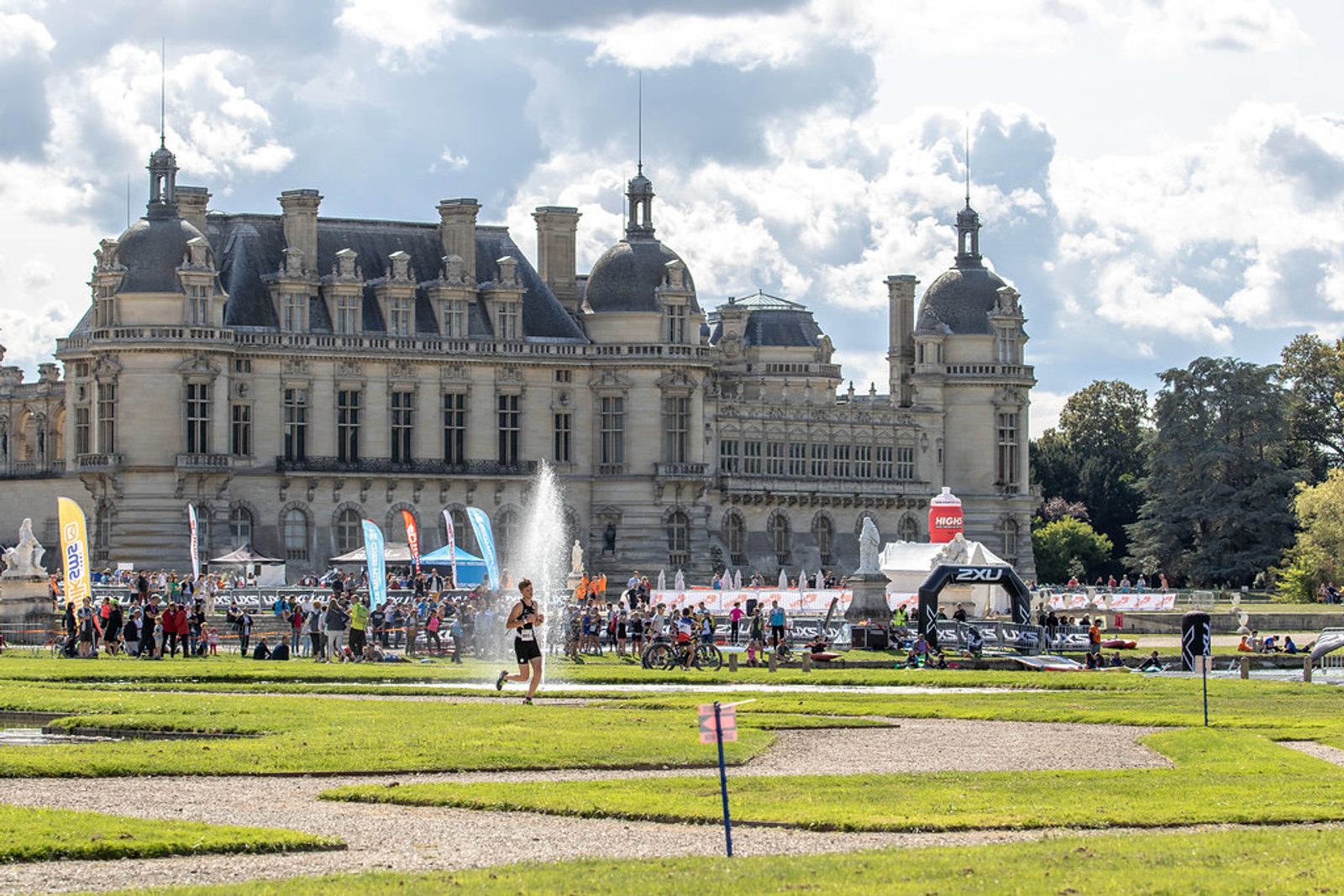 A runner is on a path in front of a large historic chateau with multiple towers and ornate architecture. A fountain is spraying water near the path, and a crowd of people is gathered with flags and tents in the background under a cloudy sky.