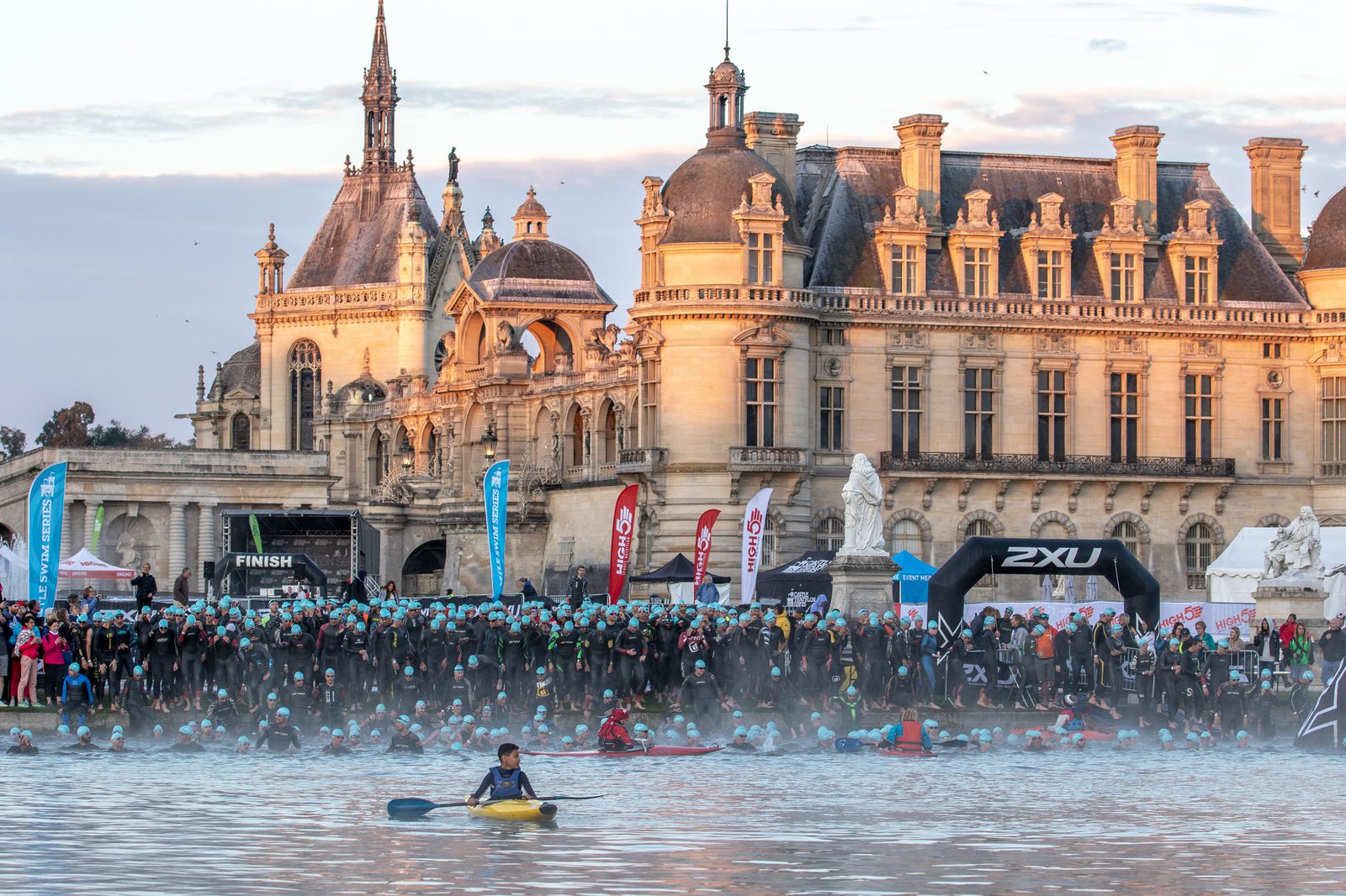 A large group of swimmers in wetsuits and caps are starting a race in a misty river. Behind them is a grand, historic chateau with intricate architecture and tall towers. Spectators and colorful flags are visible along the shore, with a kayaker in the foreground.