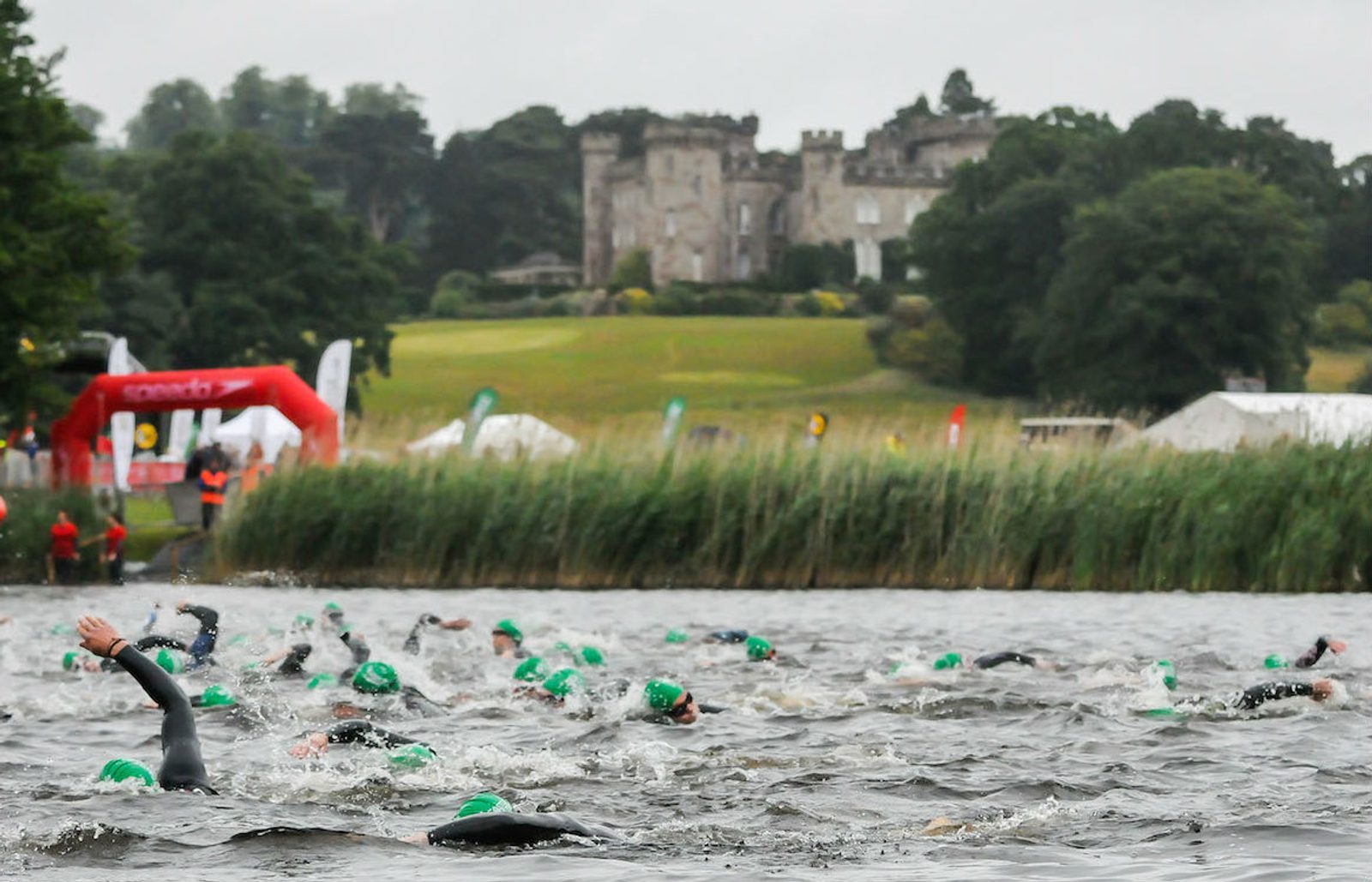 Athletes wearing green swim caps participate in an open water swim race in a lake, with a large historic building surrounded by lush greenery in the background. Marshals and tents are visible along the shoreline.