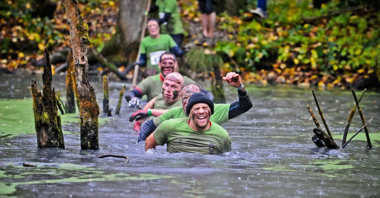 A group of people wearing green shirts and headbands wade through a muddy forested obstacle course, smiling and cheering. They navigate through water filled with sticks and tree branches. The scene is lively with determination and camaraderie.