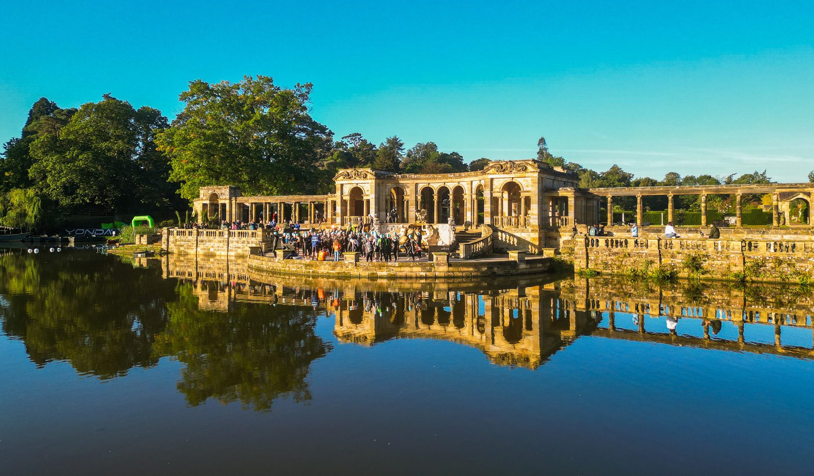 A large group of people gather in front of historic ruins with elegant columns and arches, next to a reflective pond under a clear blue sky, surrounded by lush greenery. The scene suggests a celebratory event or gathering.