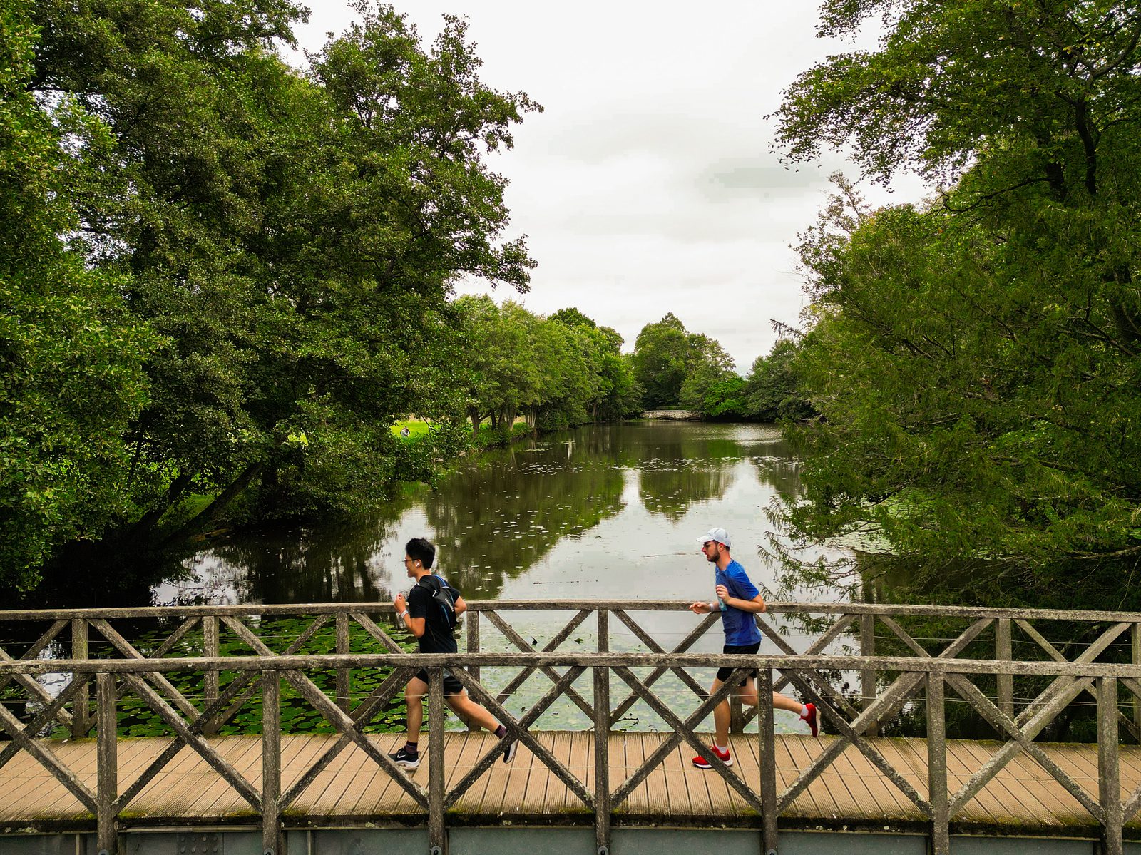 Two people are jogging on a wooden bridge over a calm river, surrounded by lush green trees and vegetation. The sky appears overcast.