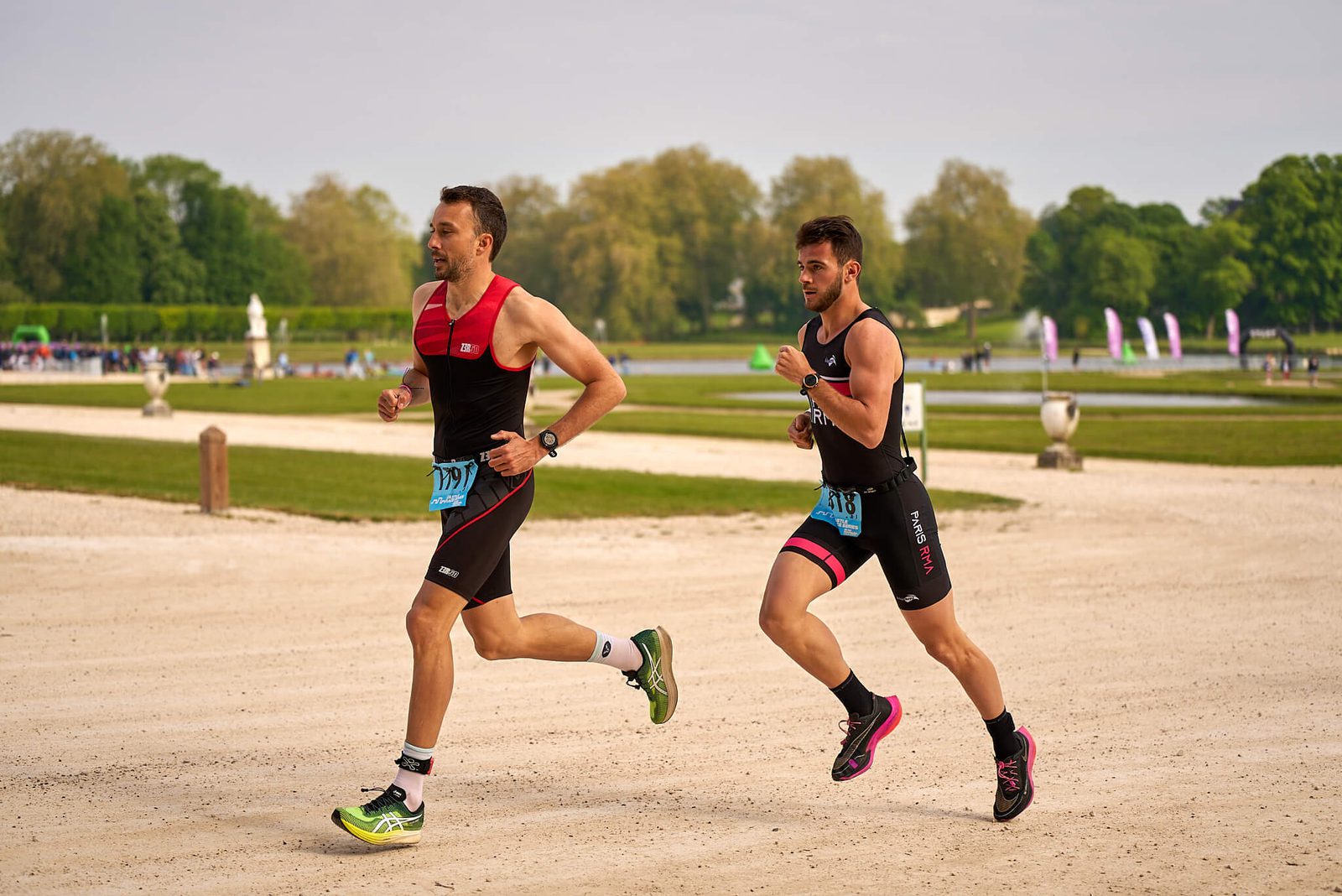 Two athletes, wearing numbered bibs, running on a gravel path in a park during a triathlon event. Both are dressed in black and red athletic gear, with one slightly ahead of the other. The background features green lawns, trees, and spectators in the distance.