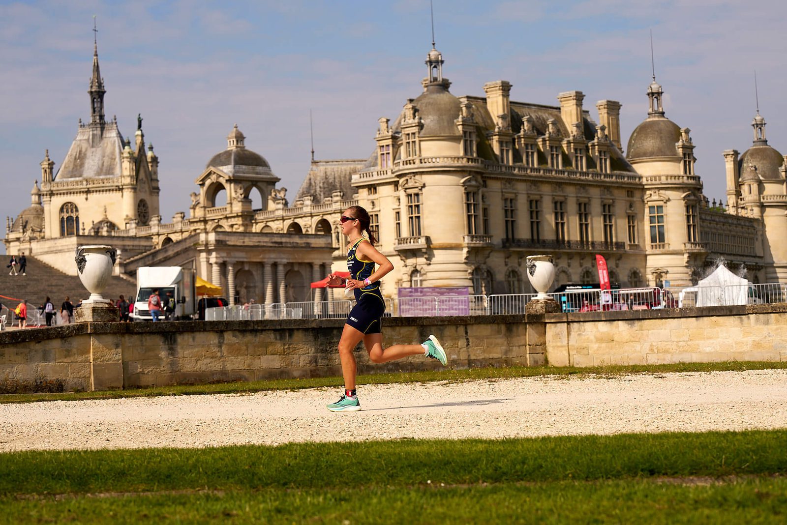 A person wearing athletic gear is running on a gravel path in front of an ornate, historic building with multiple towers and spires under a partly cloudy sky. The setting suggests a scenic location, hinting at a race or marathon taking place.