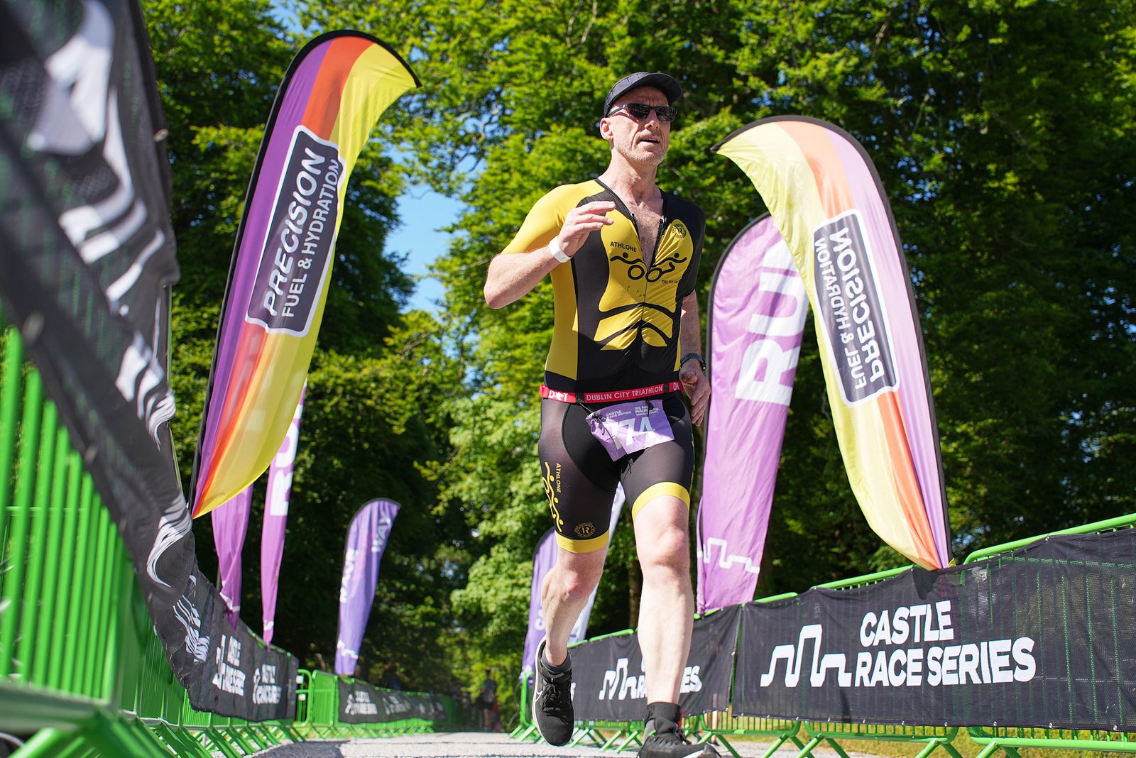 A person in athletic gear and sunglasses is running in a race, passing a set of colorful banners and green fencing. The background has lush green trees, and the banners display sponsors and "Castle Race Series" branding.
