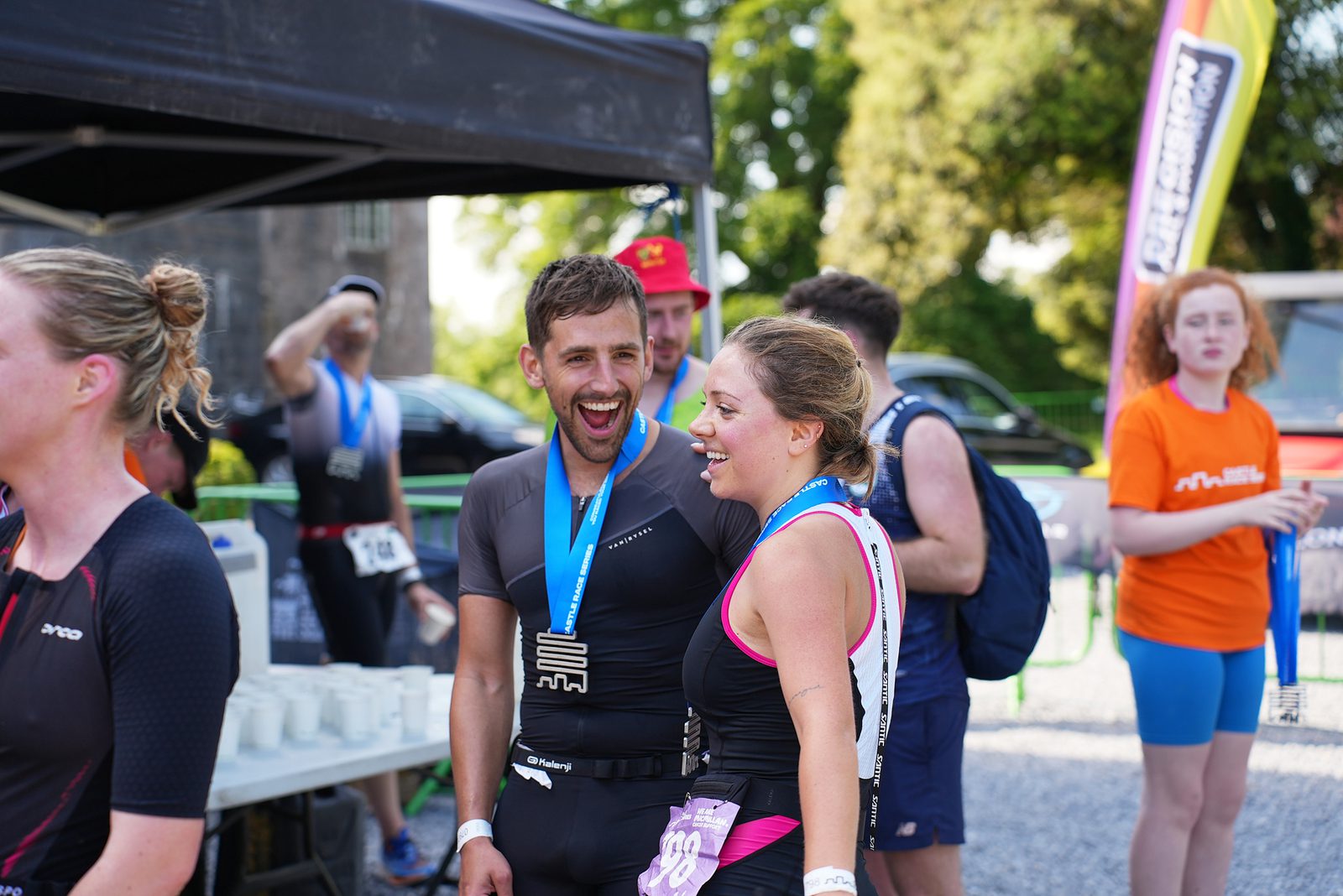 A group of people stand near a tent during an outdoor event on a sunny day. Two individuals with medals around their necks are smiling and talking. Others in the background are engaging in various activities. The scene is vibrant and filled with energy.