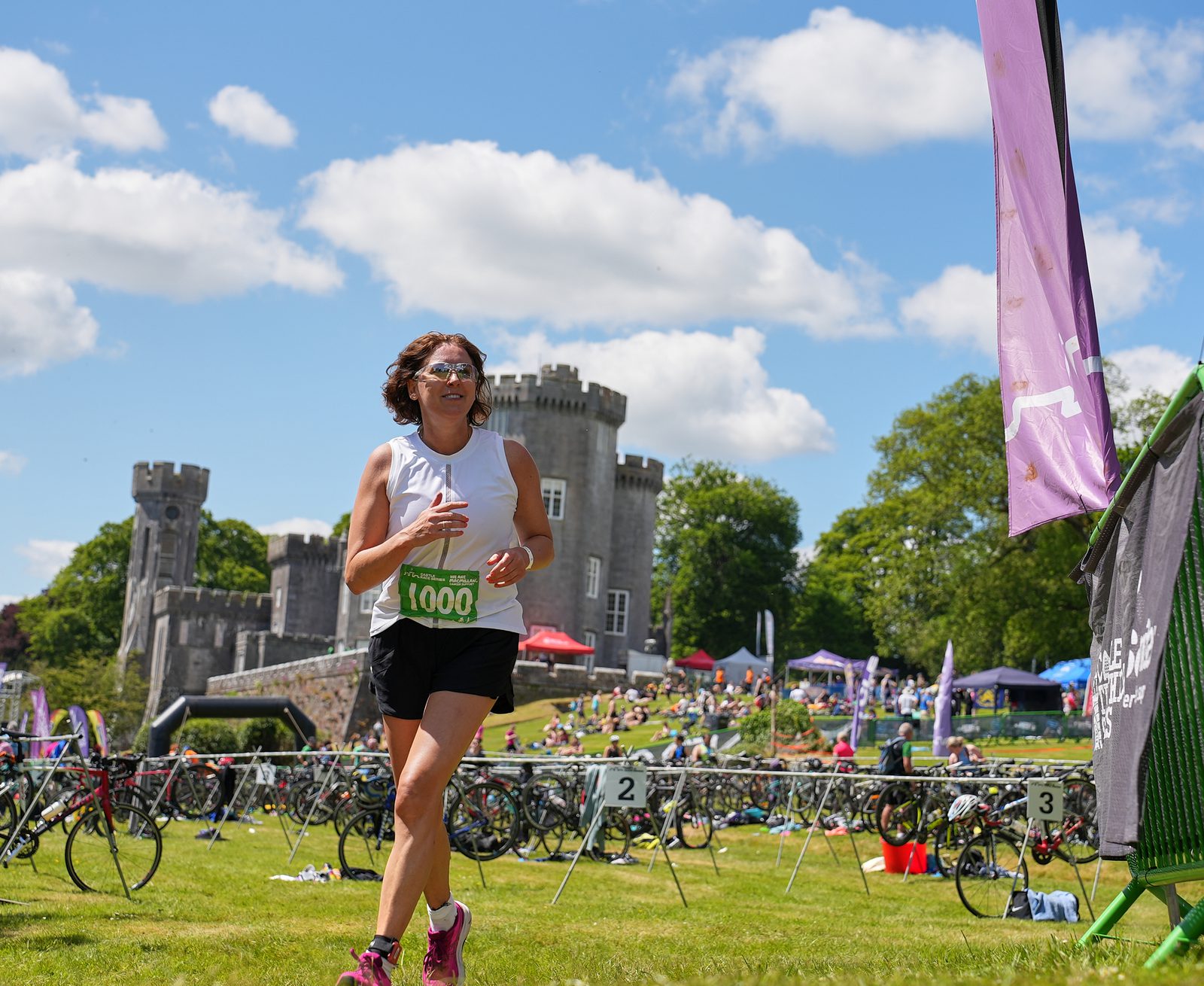 A person wearing a white tank top, black shorts, and sunglasses, with race bib number 1000, runs on a grassy field near a castle. The background features bikes, colorful tents, people relaxing, and a purple banner. It's a sunny day with scattered clouds in the sky.