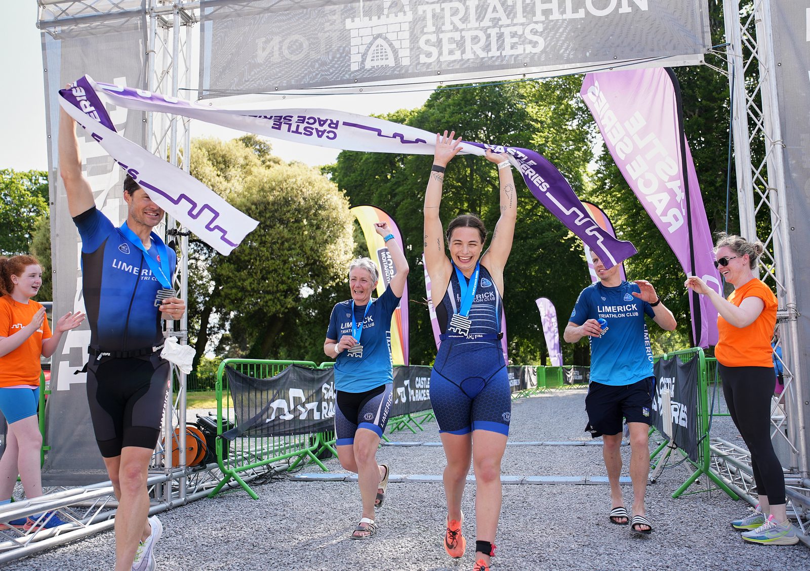 A triumphant woman crosses the finish line of a triathlon, holding the ribbon above her head in celebration. Three other participants, two men and a woman, follow closely behind, smiling and applauding. Volunteers in orange shirts stand nearby, clapping and cheering.