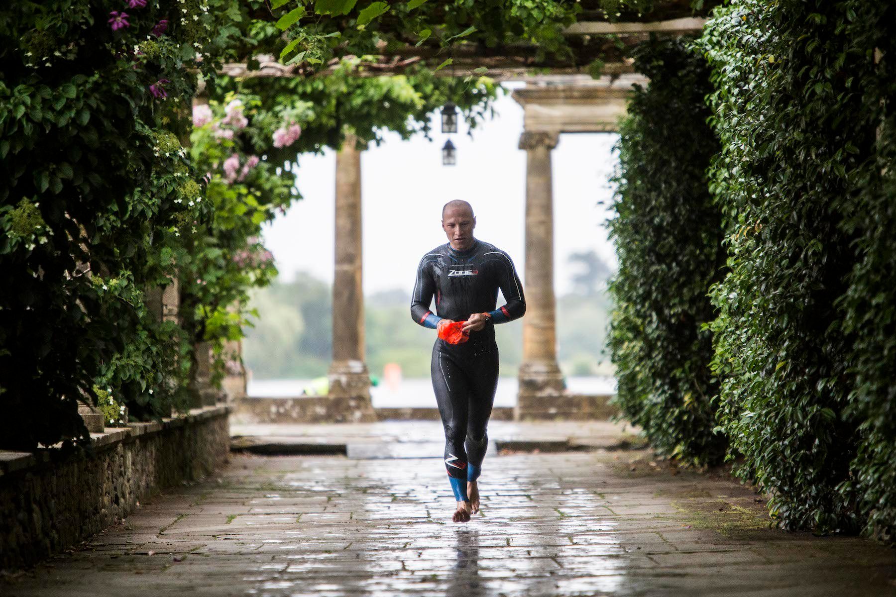 A person in a wetsuit and swim cap walks along a stone path bordered by lush greenery. The individual is barefoot and appears to be drying off with a towel. The background features an arched pergola structure with climbing plants and a view of water.