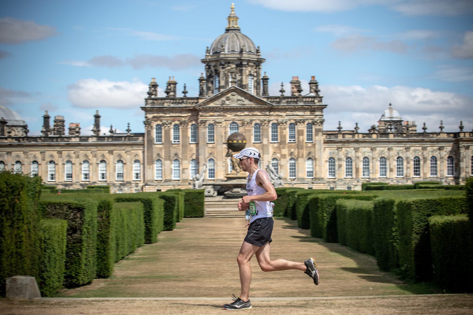 A person dressed in athletic gear, including a white cap and sunglasses, is running along a manicured garden pathway with neatly trimmed hedges on both sides. In the background, a grand and ornate historical building with a large dome is visible under a partly cloudy sky.