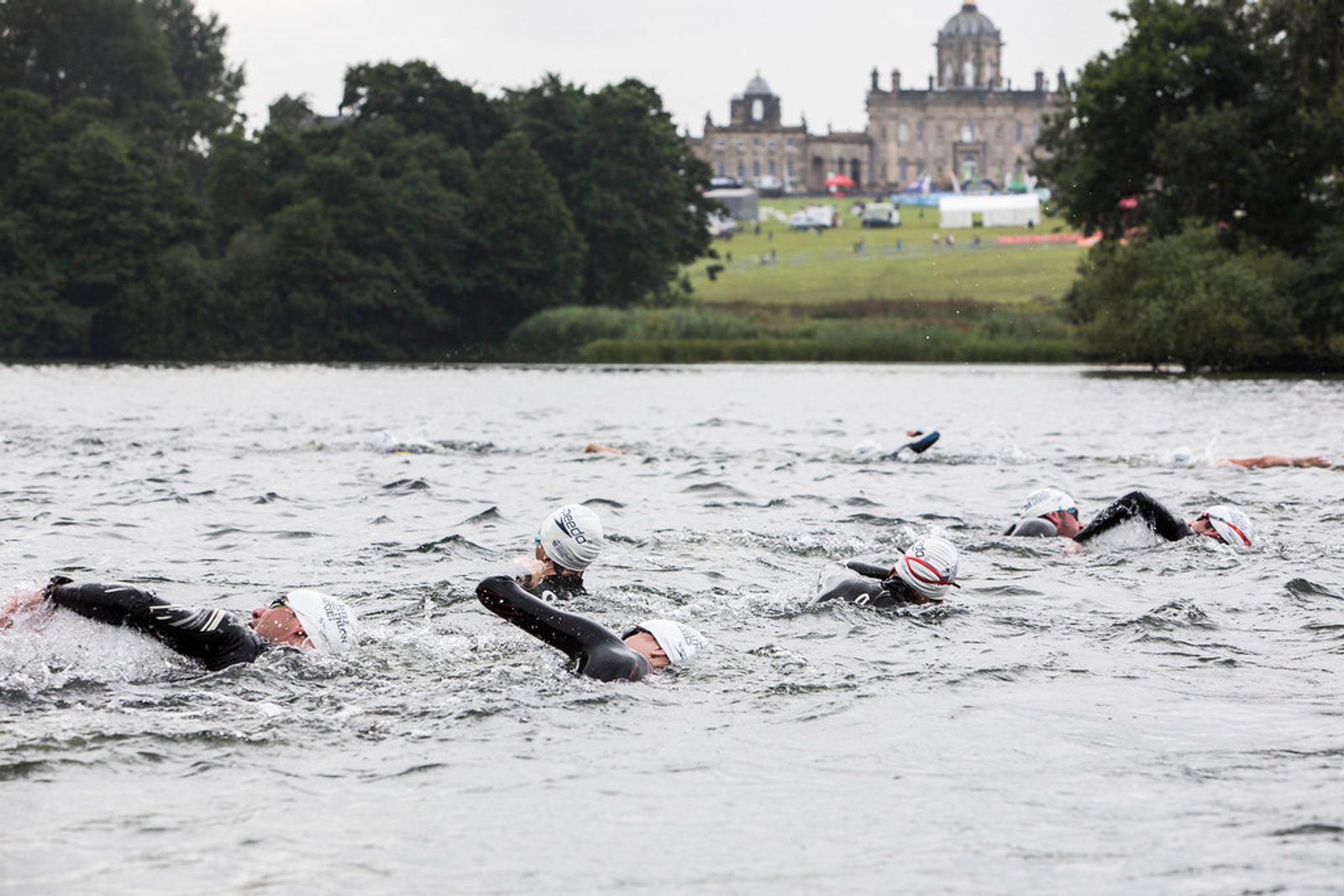 A group of swimmers in wetsuits and swim caps swim in open water during a triathlon event. The background features a large historic building on a hill with lush green trees surrounding the area. The sky is overcast.