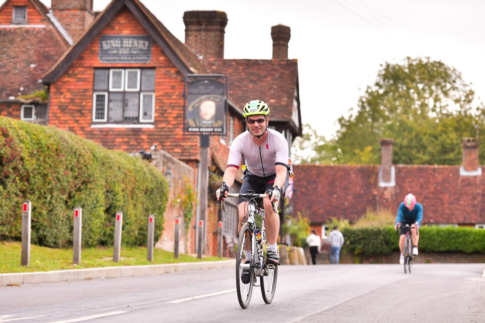 A cyclist in a white outfit and green helmet rides a bicycle on a road, smiling. Another cyclist in blue is in the background. A red-brick pub with a sign reading "King Henry" and greenery lines the street. The sky is overcast, suggesting a cool day.