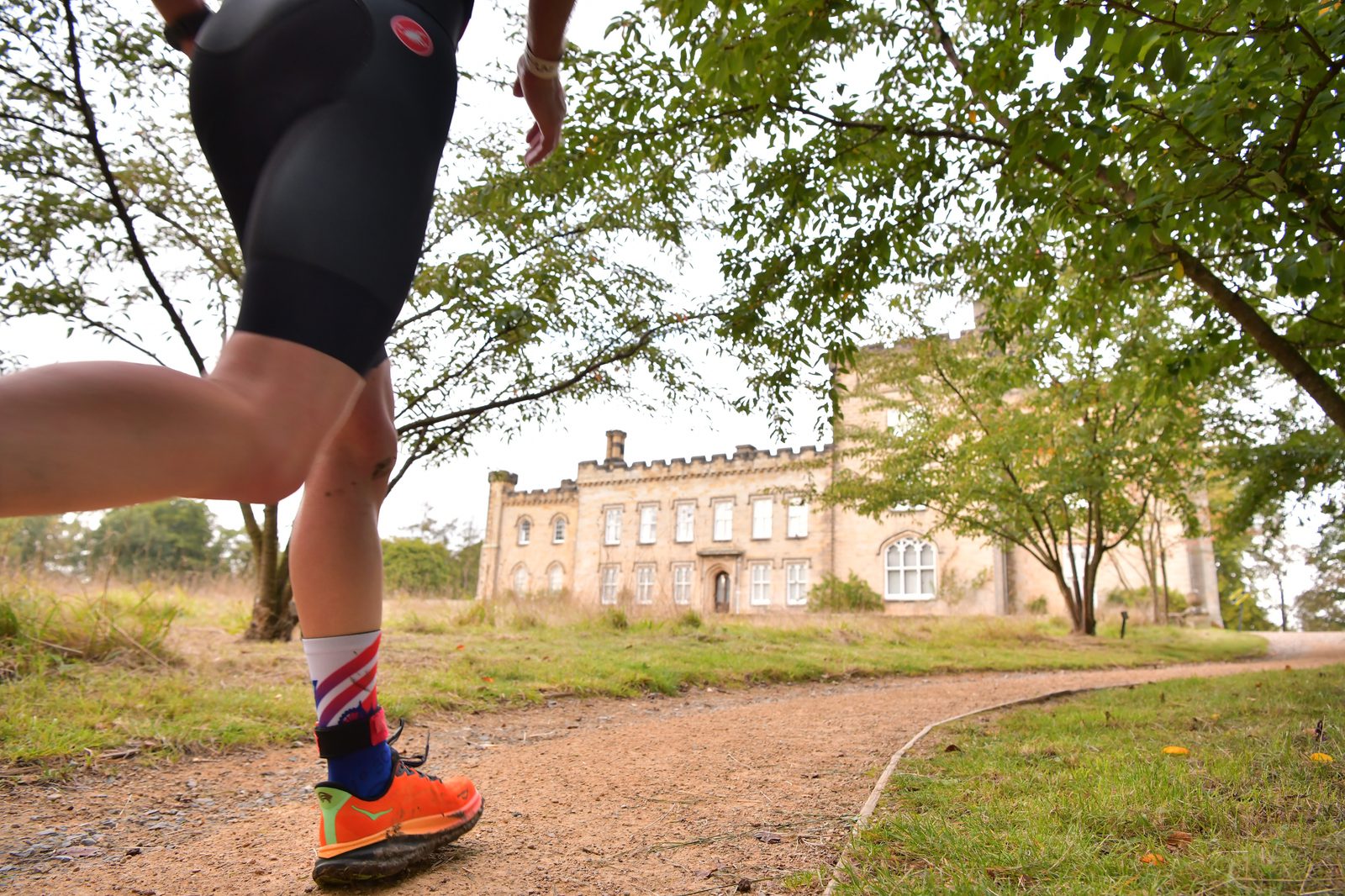 A person running on a dirt path towards a large, historic building surrounded by greenery. The runner is wearing black shorts, orange shoes, and a colorful sock. The building has a beige, stone facade with multiple windows and a crenellated roofline.