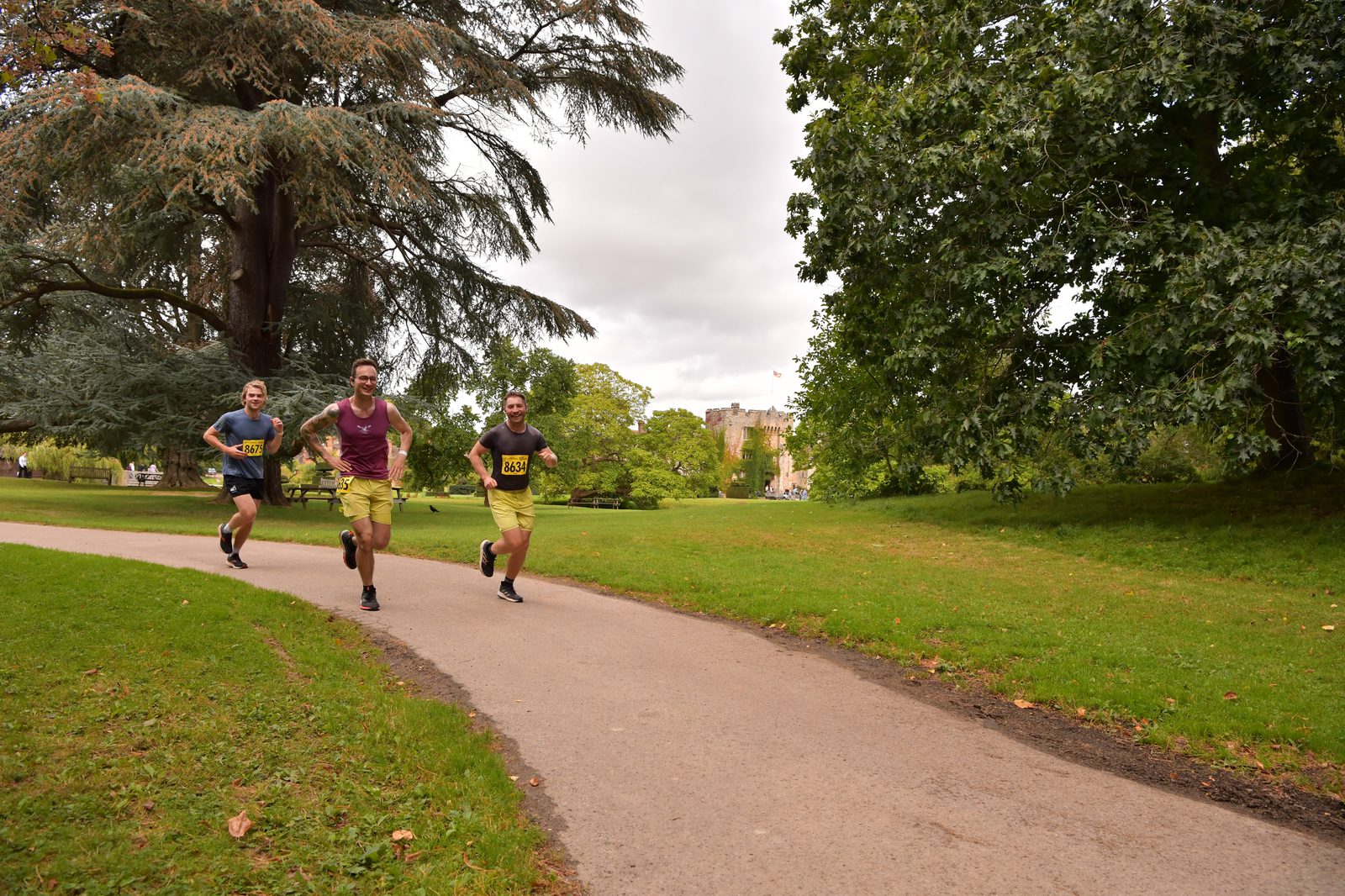 Three runners jog along a paved path through a lush green park. Two men wear numbered race bibs, and another runner follows closely behind. Tall trees and grass surround the scene, with a distant stone building visible in the background under a cloudy sky.
