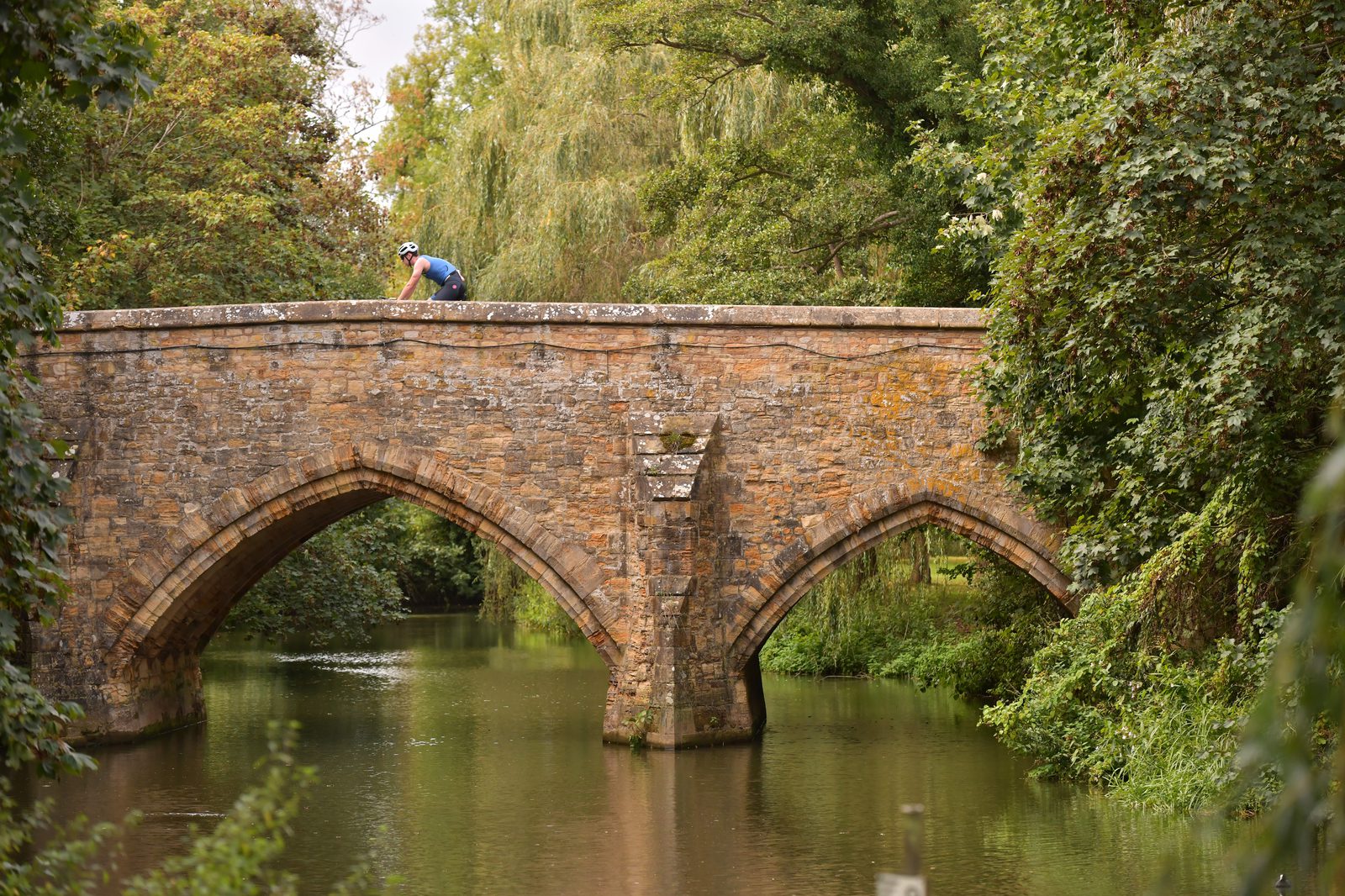 A person wearing a helmet and cycling gear rides a bicycle across a historic stone bridge over a calm river, surrounded by lush green trees and foliage. The bridge features two large arches and appears to be aged and weathered.