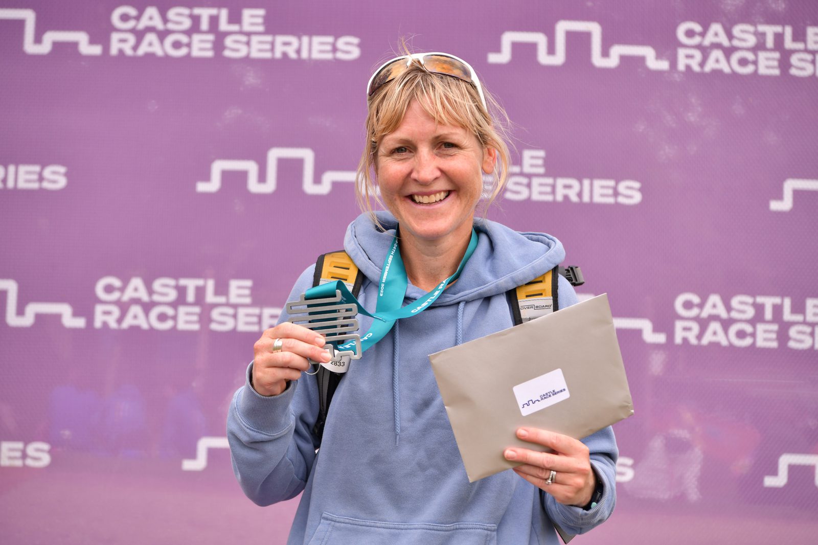 A smiling person wearing a light blue hoodie and sunglasses on their head holds a medal and an envelope against a purple backdrop that reads "CASTLE RACE SERIES" several times. The person is also wearing a blue lanyard around their neck.