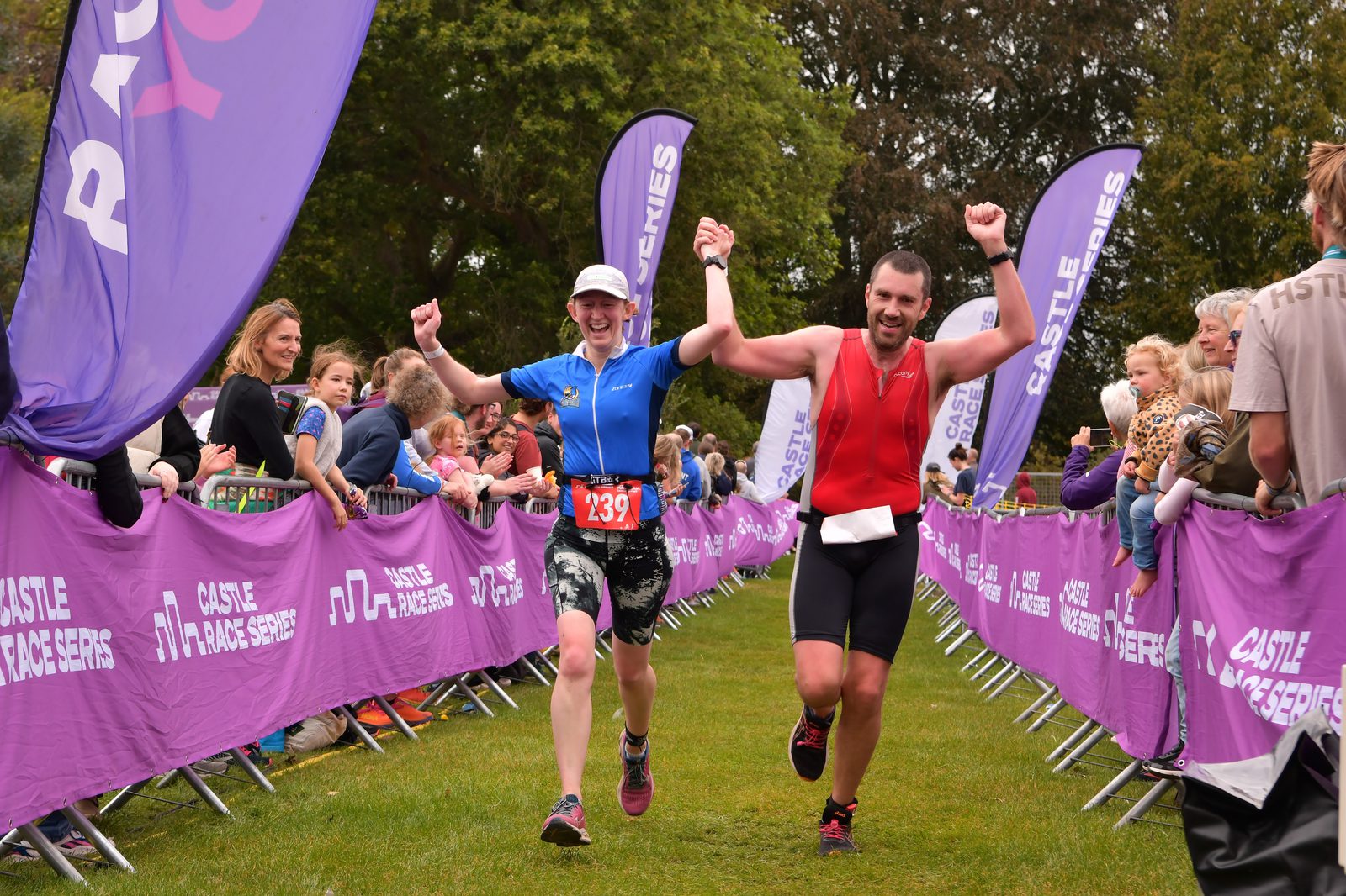 Two athletes, a woman in a blue shirt and a man in a red top, celebrate as they cross the finish line of a race, holding hands and raising their arms in triumph. Spectators stand behind purple barriers with "Castle Race Series" banners. Trees are visible in the background.