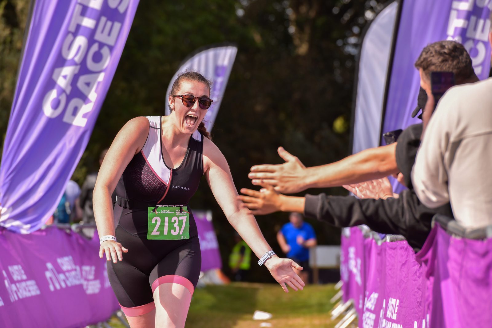 A female athlete with race bib number 2137 smiles enthusiastically while running and giving high-fives to supporters. She wears sunglasses and a black and pink athletic outfit. Purple banners reading "Castle Races" line the path in the background.